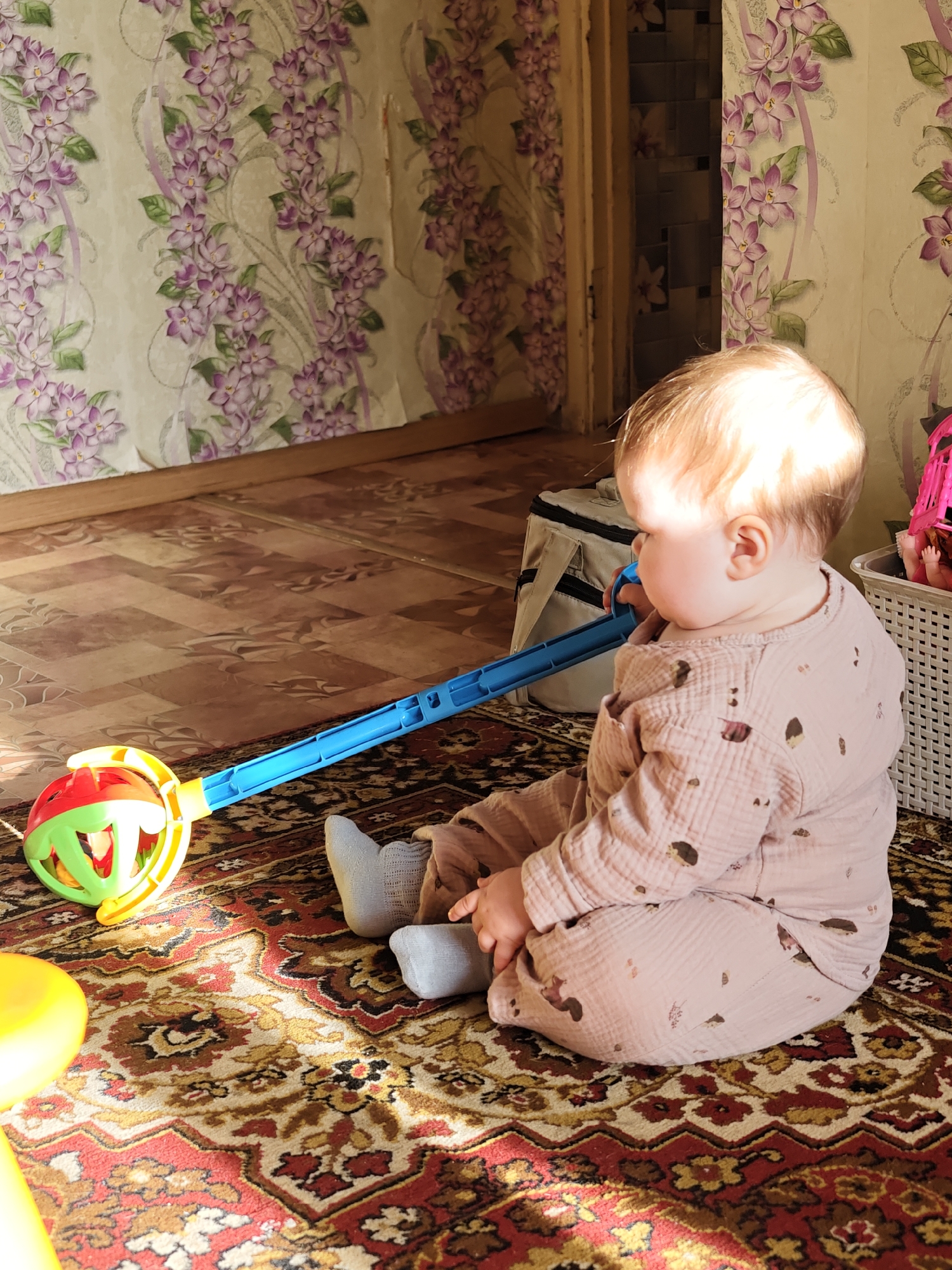 A baby sits on a rug in a room with floral wallpaper, potentially in the city of Уральское, Russia. The baby is wearing a patterned pink outfit and gray socks. A blue toy with a colorful ball end is next to the baby on the rug. The rug has a complex pattern of red, gold, and brown. A diaper bag and toy basket are visible in the background. Sunlight illuminates part of the rug and the baby's head.