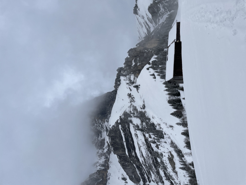 The image depicts a winter mountain landscape in Passy, France, under an overcast sky. In the foreground, a vast expanse of fresh snow covers the terrain, featuring a faint trail of footprints originating from the right and moving towards the lower center. In the midground, two dark-brown wooden structures, resembling cabins or sheds, stand on a snowy slope; their roofs are uniformly covered in snow. A sparse line of snow-dusted evergreen trees extends behind these structures and ascends the lower mountain slopes. The background is dominated by rugged, snow-covered mountains with visible rock formations, partially obscured by low-hanging grey clouds or fog that merge with the overcast sky. The overall lighting is diffused, indicating a cloudy day. No individuals or active subjects are visible.