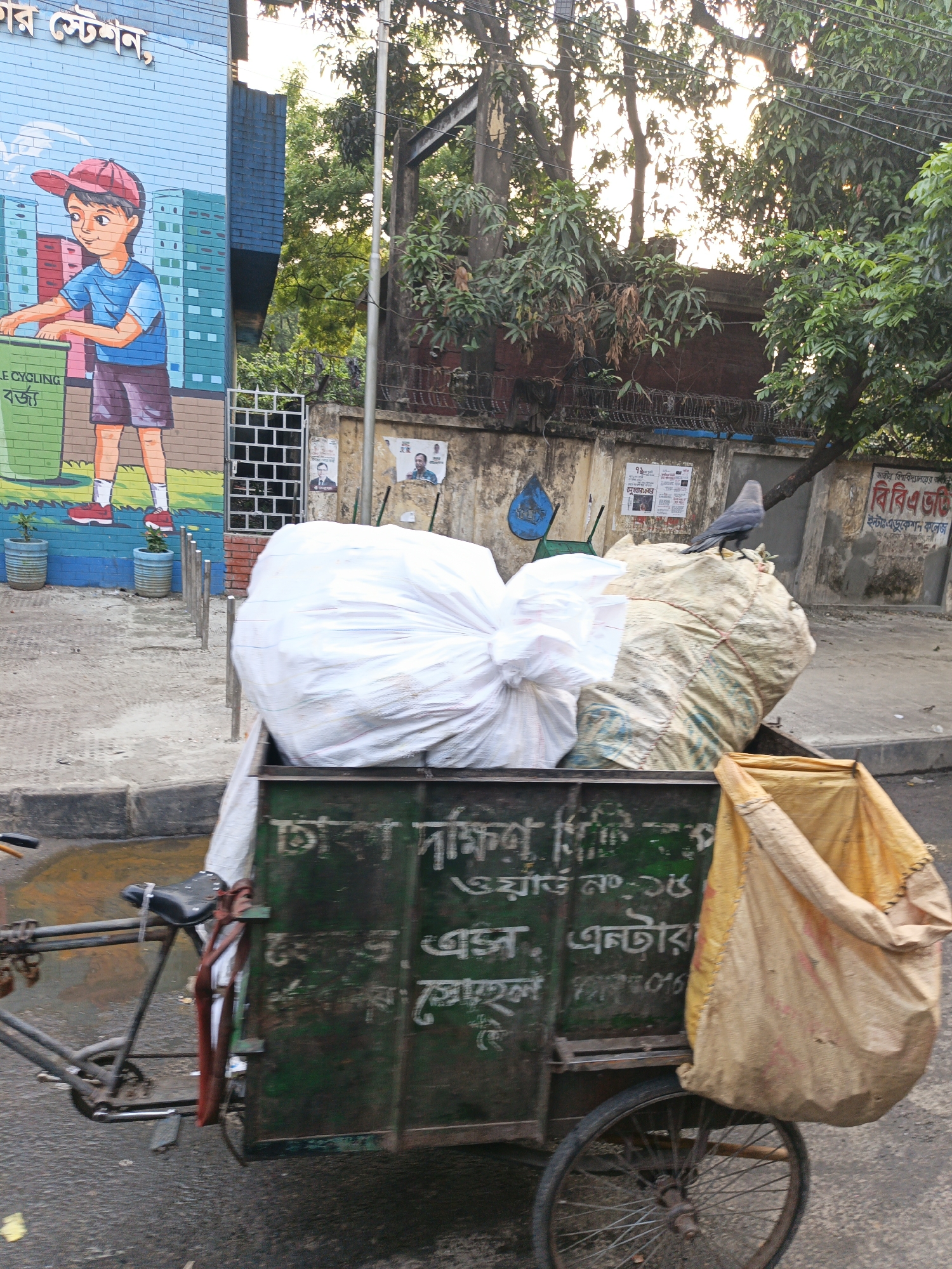 A medium shot shows a cart pulled by a bicycle filled with trash, photographed in an urban setting. The cart is dark green and contains a large white bag, a beige bag with visible seams, and a yellow cloth bag hanging off the side. The cart's side panel has faded white Bengali text. A dark gray bird stands atop the beige bag. The bicycle attached to the cart has a black seat and a visible tire. To the left, a mural depicts a young boy in a red hat and blue shirt placing trash into a green bin marked "Recycling." Next to the mural, small potted plants sit on the ground. The background includes a building with a brick façade and a wrought iron gate, along with posters and trees. The sky is bright, and the image is taken during daylight.