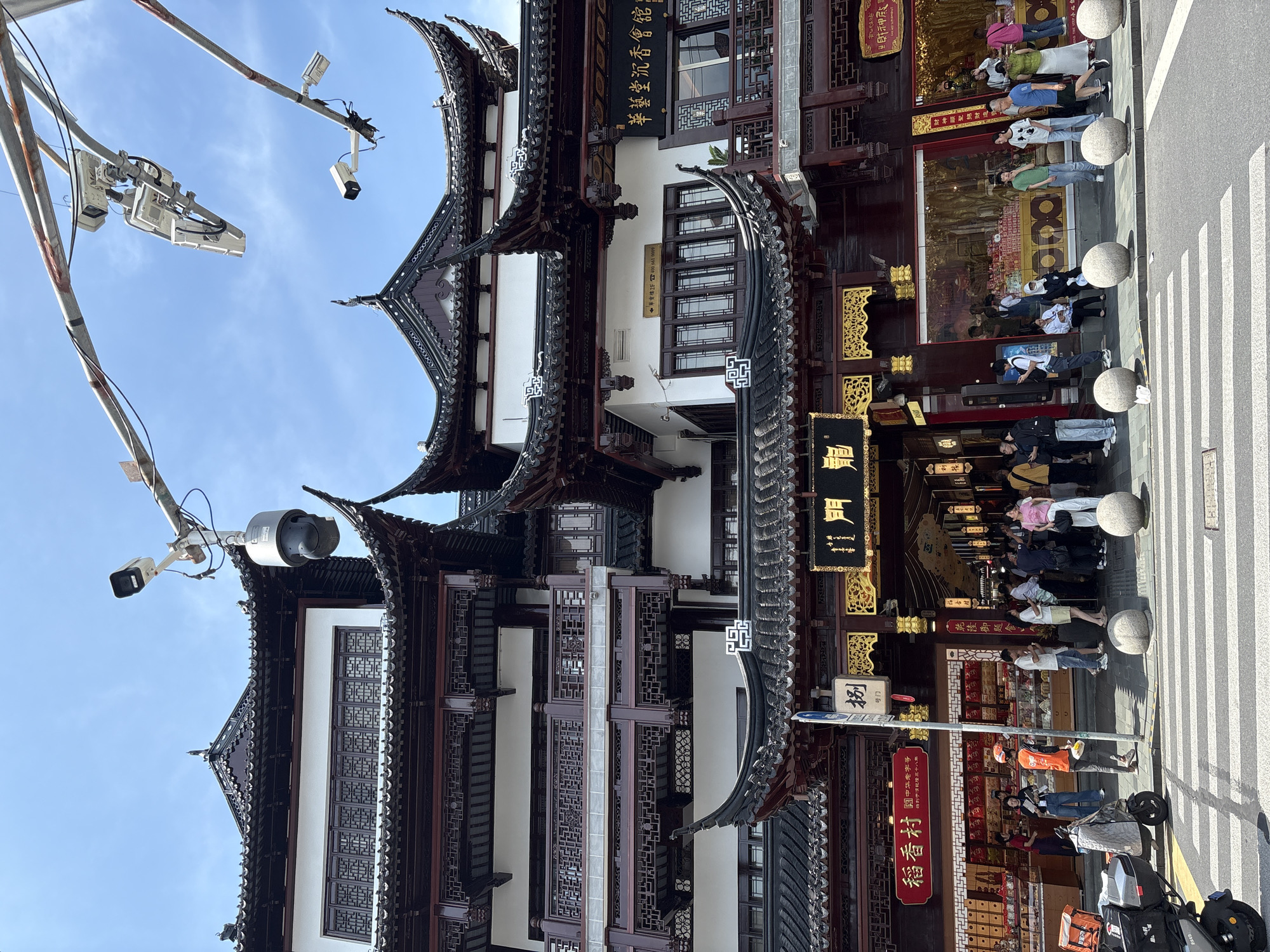 The image shows a street scene in Shanghai, China, likely within a historic district given the architecture. In the foreground, a crosswalk with white stripes is visible on a paved road. Several round, grey stone bollards are positioned along the sidewalk. To the left of the crosswalk, a black electric scooter with orange storage containers is parked.
Dominating the background is a multi-story building with traditional Chinese architecture, characterized by dark tiled, curved roofs with ornate details. The building's facade features white walls and dark wooden accents, with intricately patterned windows. Various signs with Chinese characters are displayed on the building and its storefronts. One prominent sign above an arched entrance reads "豫园商城" which translates to "Yu Garden Shopping Mall". Another red sign on the left says "稻香村" (Dao Xiang Cun), a well-known pastry shop.
A crowd of people, dressed in casual clothing, are gathered in front of the entrance and along the sidewalk, some appearing to be shopping or waiting. Several individuals are looking at their phones, and others are engaged in conversation. A food delivery person wearing an orange vest and helmet is standing near a store. Above the building, multiple security cameras are mounted on poles against a bright blue sky with scattered clouds. The overall scene depicts a busy commercial area in Shanghai.