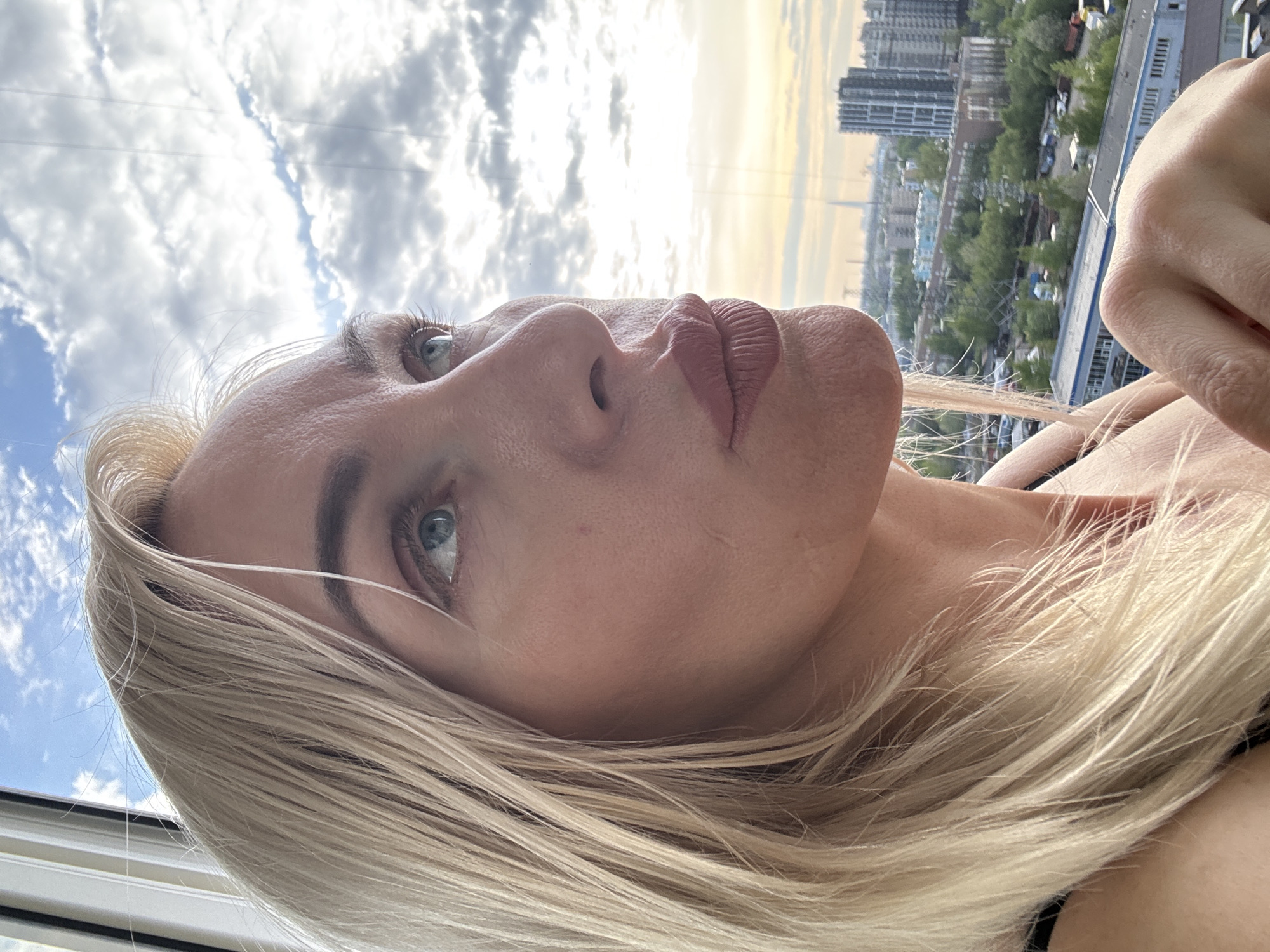 The image shows a low-angle portrait of a fair-skinned woman with blonde hair. Her face is angled upwards towards a sky filled with clouds. The background reveals an urban environment with high-rise buildings and trees, suggesting a cityscape in an unknown location. The woman's eyes are open, and she is wearing lipstick. Her hand is visible in the upper right corner of the frame.