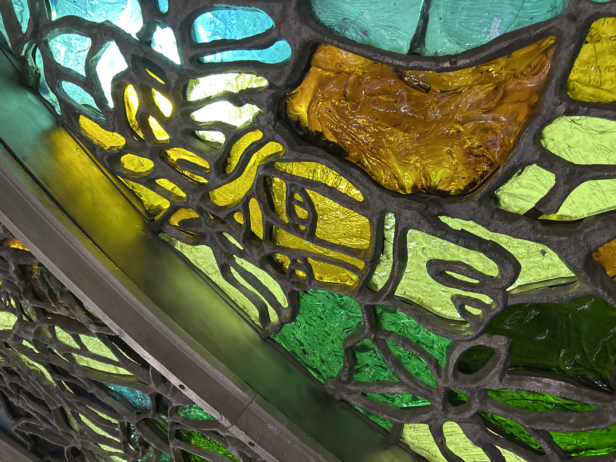 The image shows a close-up of a stained-glass window. The glass pieces are irregular in shape and are held together by a grey, branching material. The colors of the glass range from blue at the top left, through yellow and gold, to green at the bottom. Light is visible through some of the glass pieces. The scene is located in Unknown, Unknown.