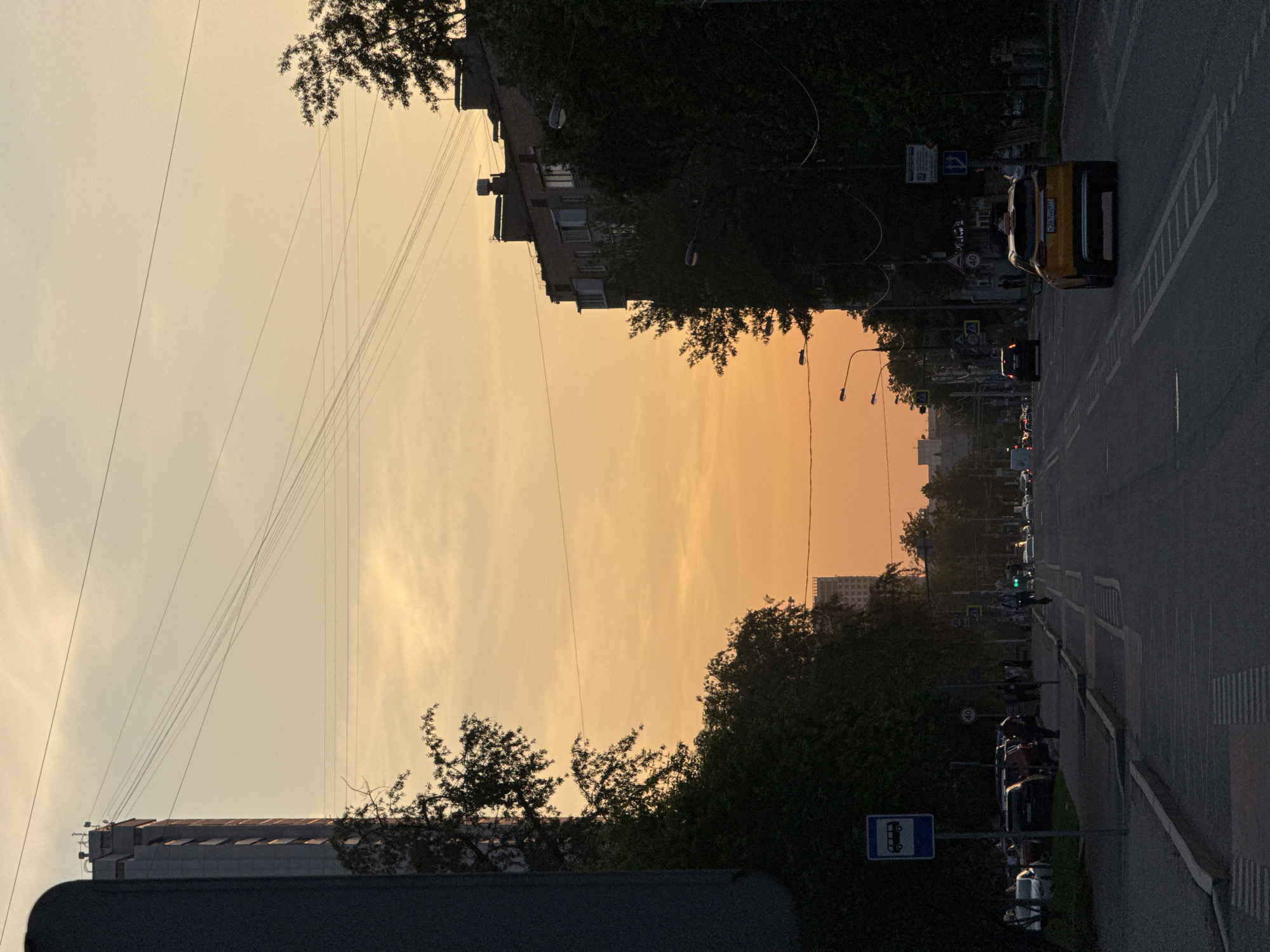 The image depicts a street scene in Moscow, Russia, at dusk or dawn. The sky is a pale orange. Buildings flank either side of a wide road, with dense trees. Power lines crisscross overhead. On the right side of the road, there is a yellow car and other vehicles. Pedestrians can be seen along the sidewalk. A bus stop sign is visible. In the distance, there are more buildings and streetlights along the road.