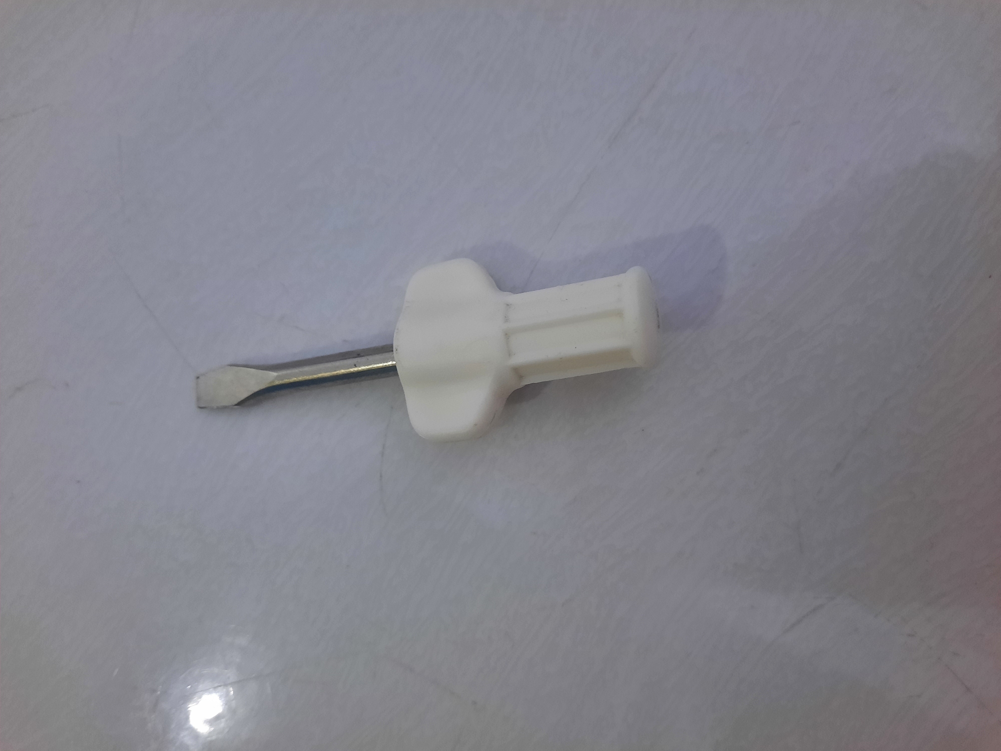 A flathead screwdriver with a white handle lies on a textured, light-colored surface. The screwdriver is positioned diagonally across the frame, with the metal tip pointing towards the upper left corner and the handle extending towards the lower right. The handle has a ribbed design. The surface appears to be a countertop or floor with subtle lines and variations in shading, possibly due to lighting or the material itself. The background is largely out of focus, emphasizing the screwdriver as the primary subject. The image does not provide any information about a location in Lhokseumawe, Indonesia.