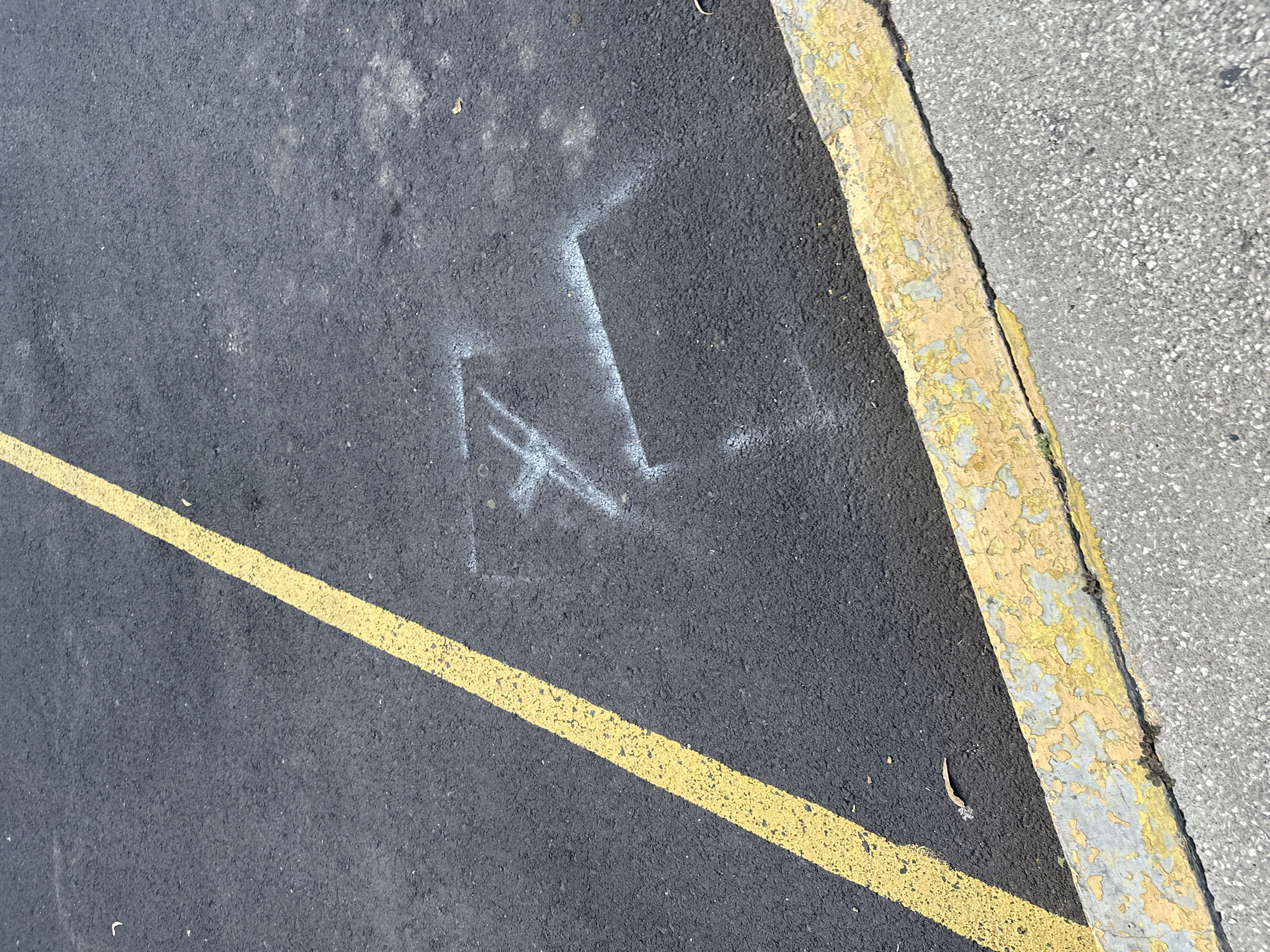 The scene shows a section of asphalt pavement. A yellow painted line marks a parking space boundary. Adjacent to the asphalt is a concrete curb painted with faded yellow paint. A white painted mark, resembling a stylized "A" with an additional horizontal line, is on the asphalt near the curb. The background consists of a section of adjacent concrete pavement. The location is unknown.
