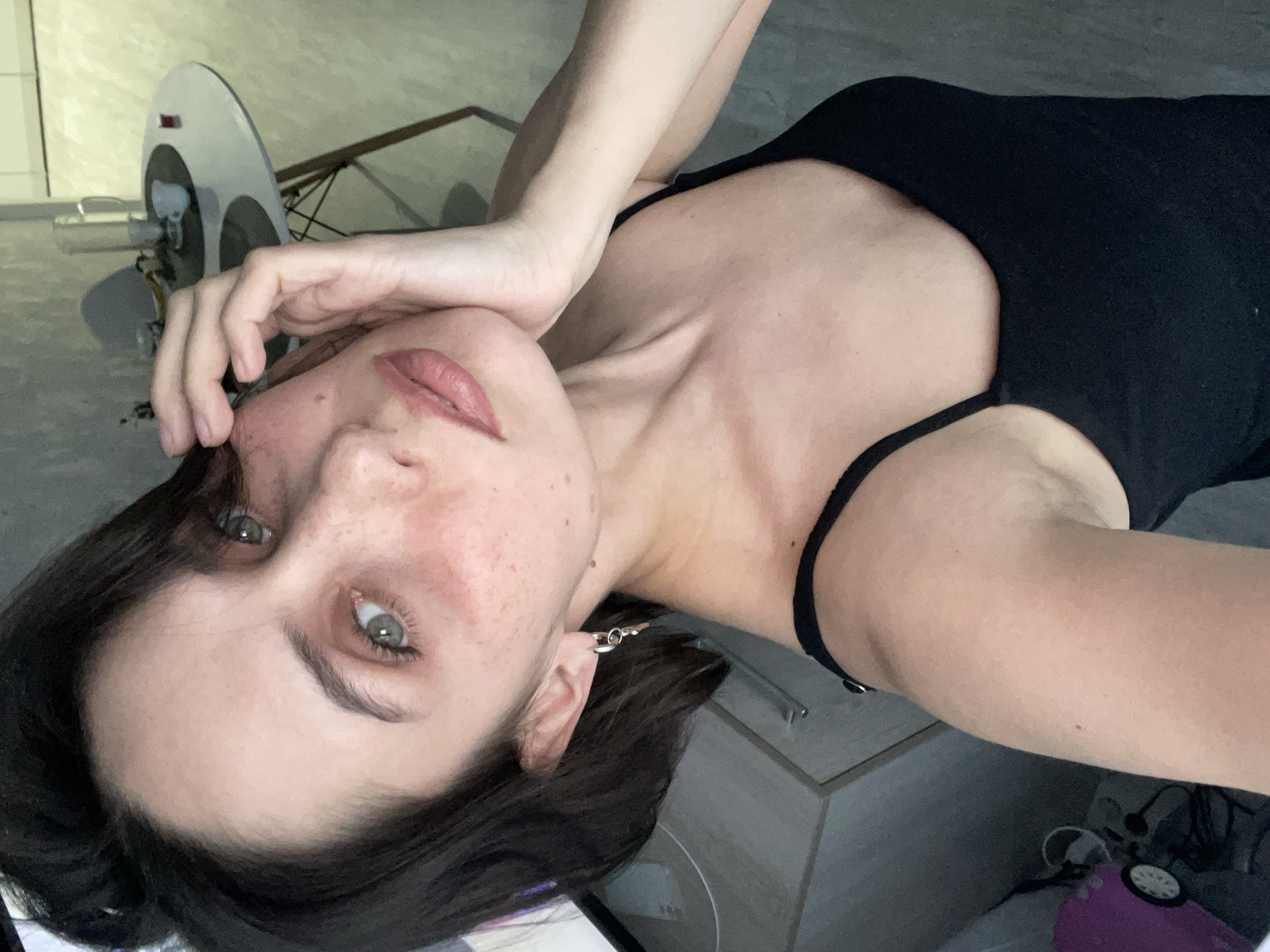 A close-up, high-angle selfie captures a young woman lying down and looking directly at the camera. She has dark brown hair, light green-grey eyes, and fair skin with noticeable freckles across her nose and cheeks. Her lips are full, adorned with a pinkish-red color, and her eyebrows are dark and well-defined. She is wearing a black tank top or camisole with thin straps.
Her right hand (viewer's left) is raised, with fingers gently resting near her jawline. A silver-toned earring, featuring a small hoop and bar design, is visible on her right ear.
The background is softly blurred but indicates an indoor setting, likely a room in a residence located in Izhevsk, Russia. A light-colored wall or floor occupies much of the background. To the upper left, behind her head, a light-colored, possibly white or grey, object with circular details is visible, resembling a fan or a piece of equipment. Further back, a light-toned piece of furniture, possibly a dresser or cabinet, can be discerned, along with hints of other household items and a purple object on the floor. The lighting is soft and appears to be natural daylight. No visible text is present in the image. Her expression is calm and direct.