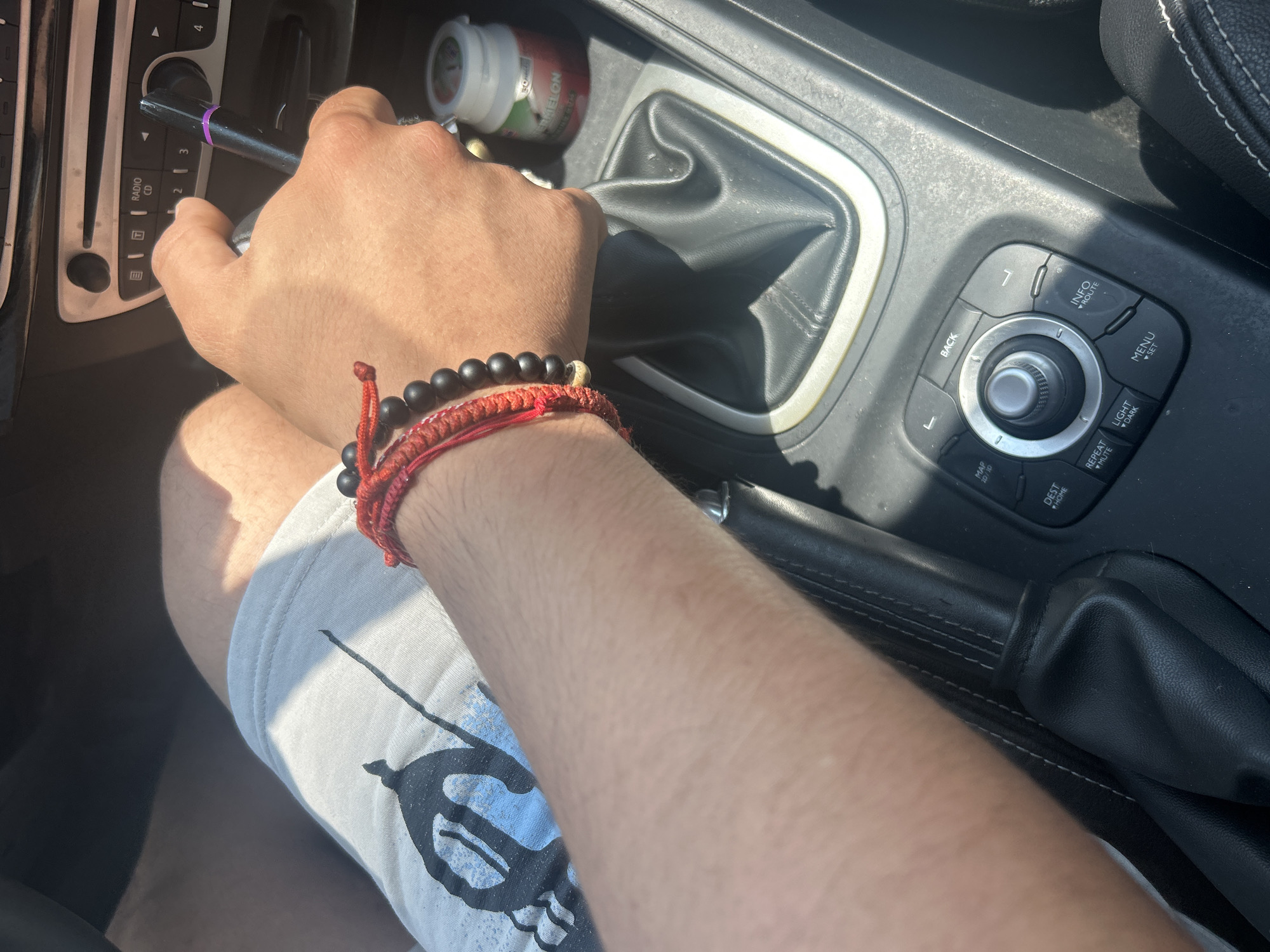 A person's right arm and hand are visible, gripping a manual gear shifter in a car. The person is wearing a red string bracelet with black beads on their wrist. They are also wearing light-colored shorts with a graphic print. The car's interior is visible, including the gear shifter, handbrake, and a portion of the center console with various buttons and a dial. A small cylindrical container with a label that includes the word "WATERMELON" is also present. The scene is lit by sunlight, suggesting it is daytime. The location is identified as being in the city of Plovdiv, Bulgaria.