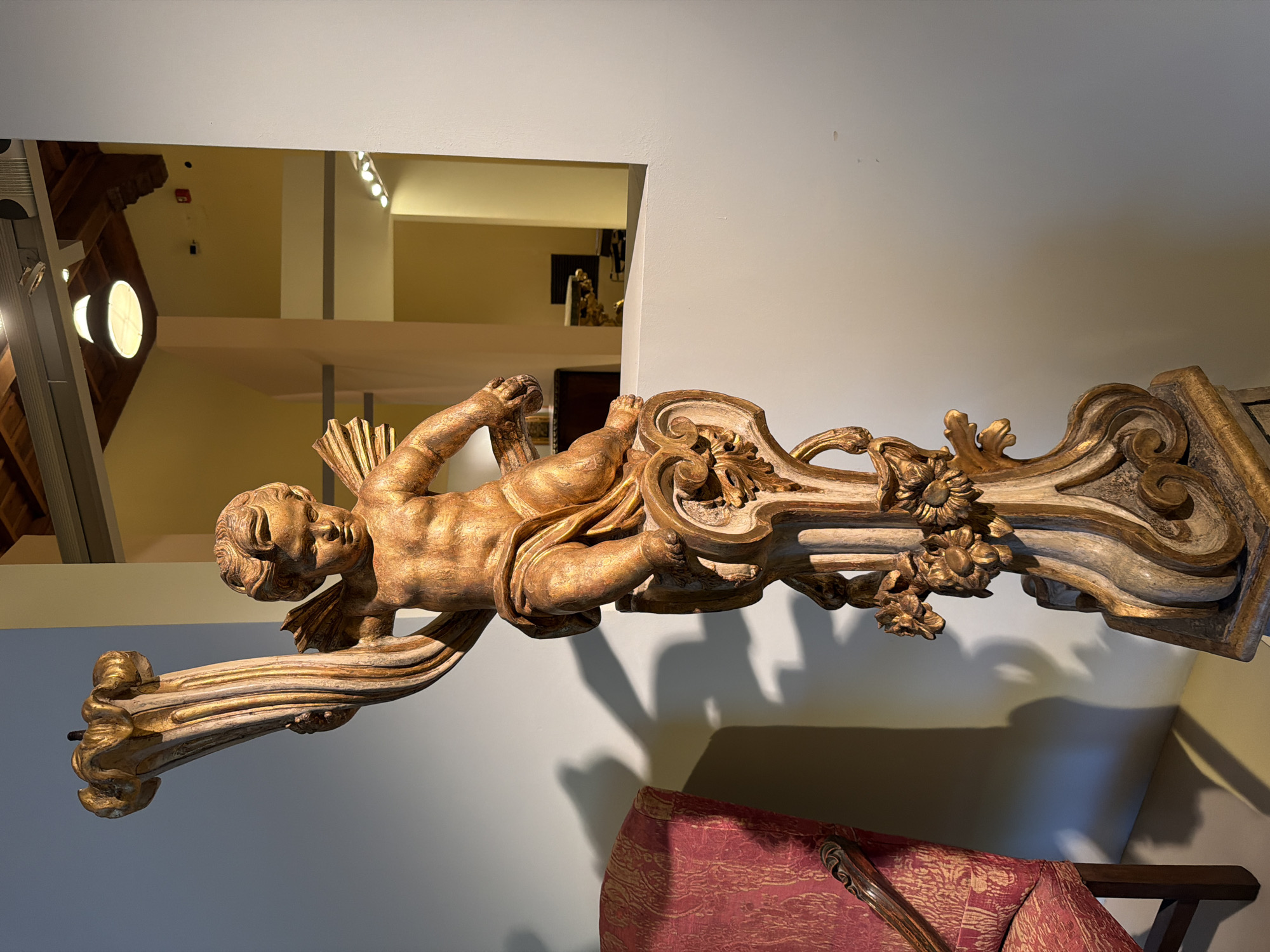 A gilded wooden sculpture of a cherub figure, likely an antique, is on display in an interior space, possibly a museum or gallery in Milan, Italy. The cherub is depicted lying on its side, with decorative scrollwork and floral embellishments surrounding it. The sculpture has a weathered appearance, indicating age. It is positioned near a wall, and a portion of a red upholstered chair is visible in the foreground. In the background, there is an architectural detail suggesting a mezzanine level or a separate room, hinting at a multi-level interior space. The lighting is a mix of natural and artificial sources, with a spotlight illuminating the sculpture.