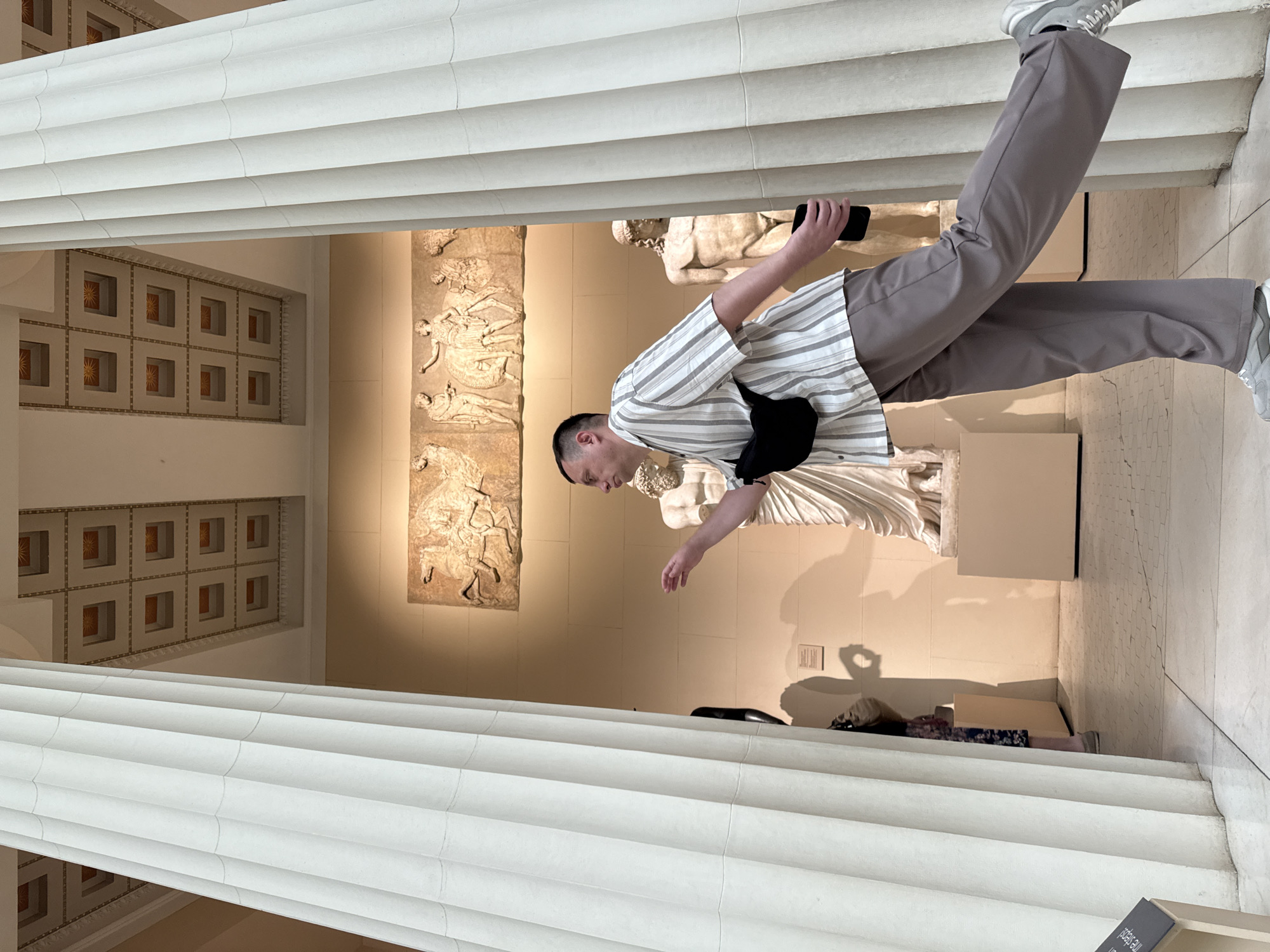 A person is seen photographing art at the Pushkin State Museum of Fine Arts in Moscow, Russia. The individual is wearing a striped shirt, gray trousers, and gray sneakers. They have a black bag slung across their body and are holding a smartphone in their right hand. The person appears to be focused on capturing an image of the statues and reliefs on display. The museum features classical architecture with detailed molding and decorative wall elements.