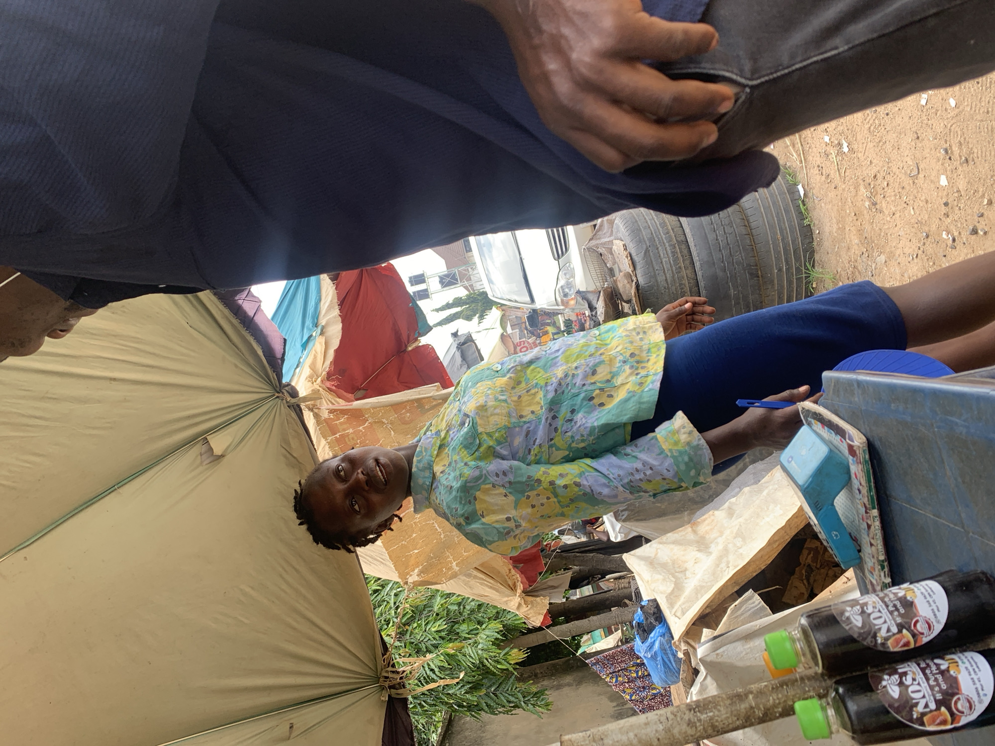 The image depicts an outdoor market scene in Abuja, Nigeria. In the foreground, a woman with braided hair, wearing a colorful floral-patterned shirt and blue pants, is looking upwards and to her right, seemingly in conversation. To her left, on a table, are two bottles of what appears to be honey, labeled "NOS Pure Organic." A blue card reader is also visible on the table. Behind the woman, a person, mostly obscured by a dark blue garment, is standing. The environment suggests a roadside stall or market with a makeshift canopy overhead, supported by poles. Stacks of tires are visible in the background, along with a white car and some hanging fabrics. The ground is sandy and appears to be unpaved.