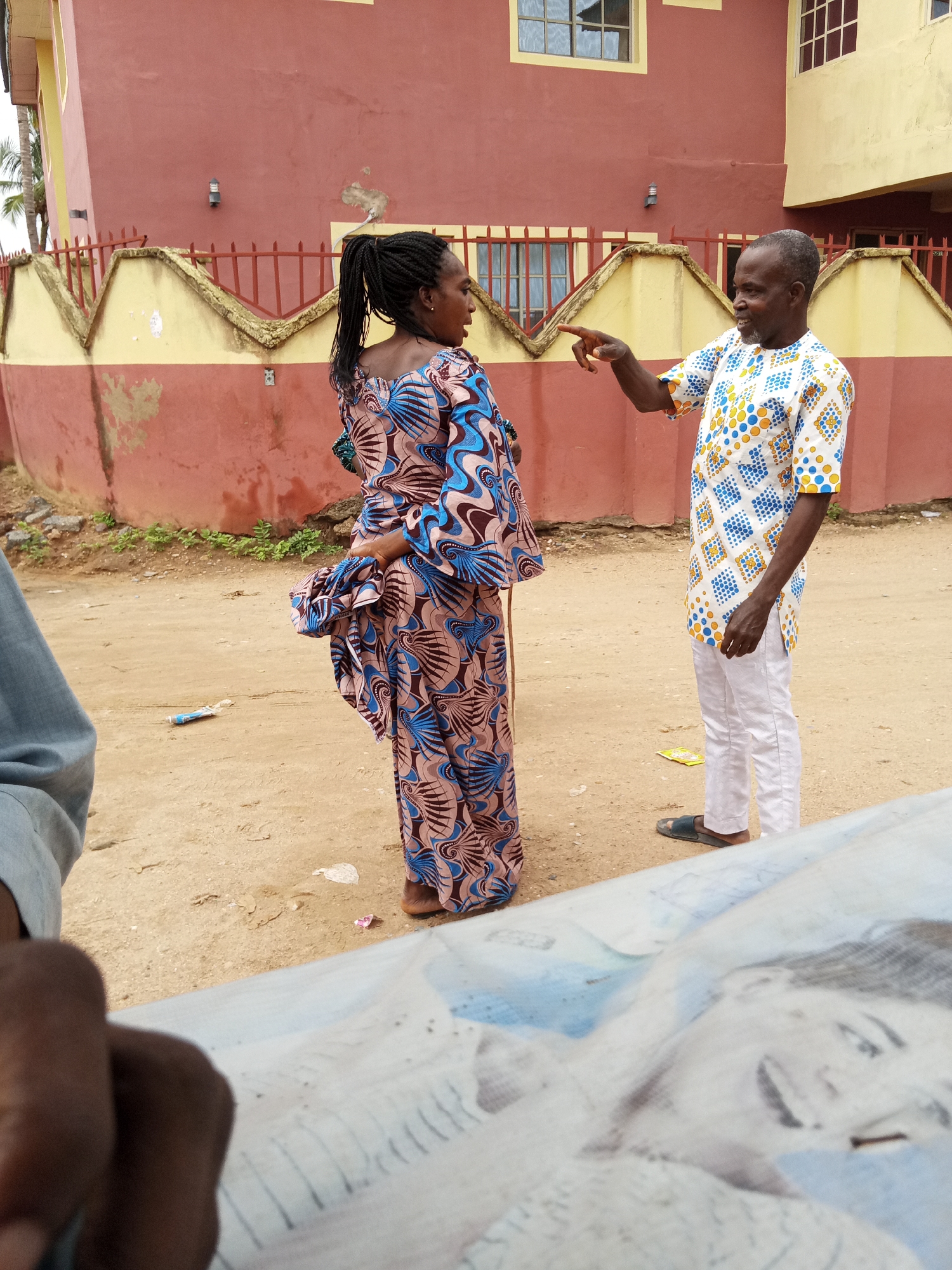 Two individuals stand on a dirt road in front of a building with a red and yellow facade. A woman with braided hair pulled back in a ponytail is dressed in a long, patterned blue and brown gown. She is facing a man who is pointing his right index finger towards her. The man is wearing a patterned white shirt with blue and yellow designs, and white trousers, and dark sandals. Litter is visible on the ground. A portion of a table or surface with a blurry pattern is in the foreground. The location is in the city of Unknown, Unknown.