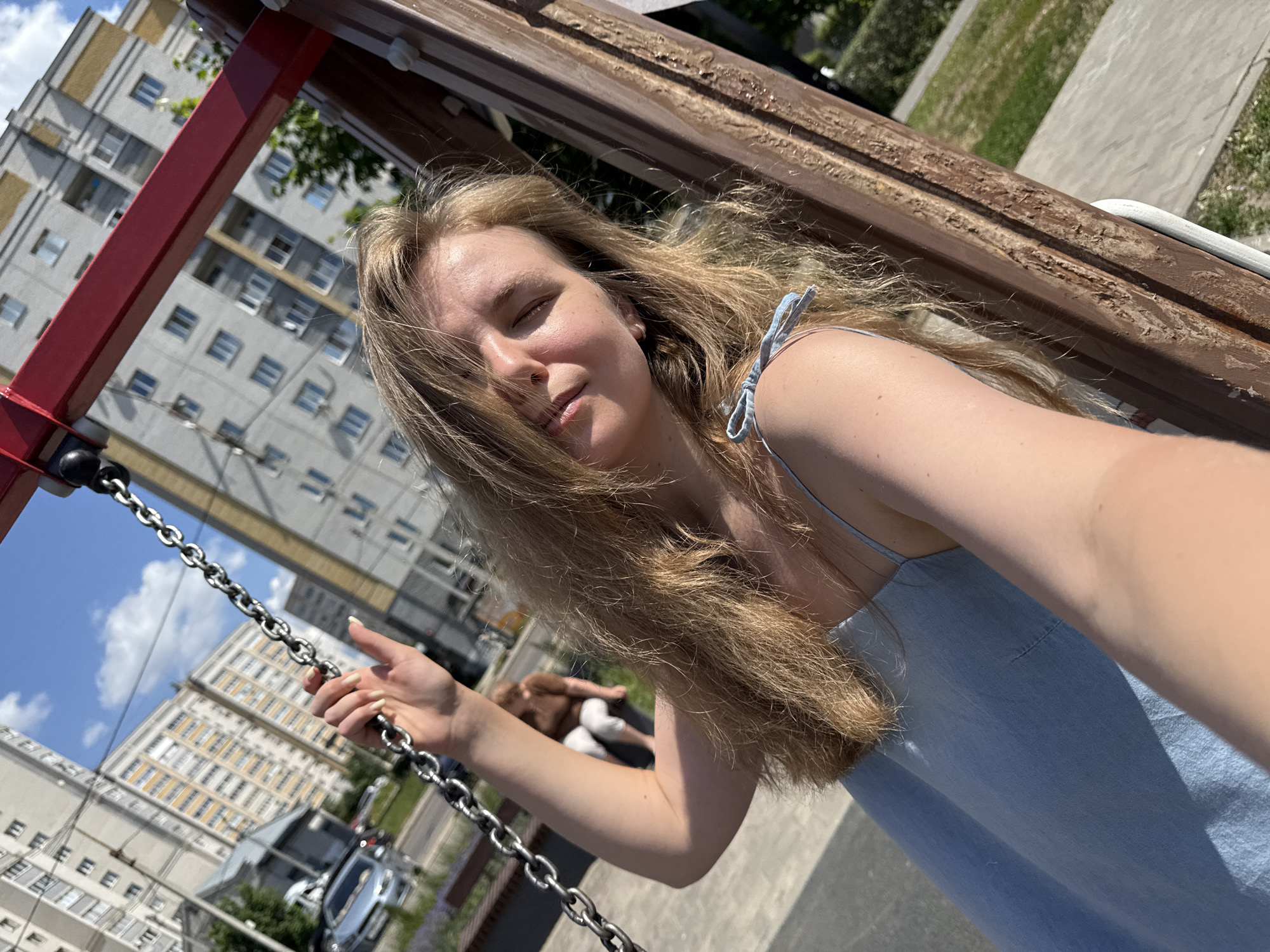 A young woman with long, blonde hair is taking a selfie while sitting on a swing. She is wearing a light blue dress with spaghetti straps. Her eyes are closed, and she is looking upwards, bathed in sunlight. Behind her, a red swing set frame is visible, along with metal chains. In the background, there are tall, multi-story apartment buildings, characteristic of urban environments. A car is parked on the street, and a person in a brown shirt and white shorts is sitting on a bench in the distance, partially obscured by the swing. The scene is set outdoors, likely in a playground or park area within a city. The presence of apartment buildings suggests an urban setting, and while the image is not specific to Vladimir, Russia, the architectural style of the buildings is consistent with that region.
