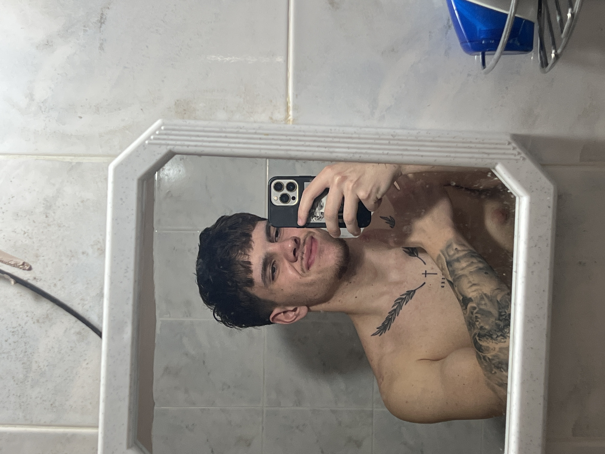 A shirtless man is taking a selfie in a bathroom mirror. The bathroom has tiled walls and a hairdryer is mounted on the wall. There is no indication of location in Sofia, Bulgaria.