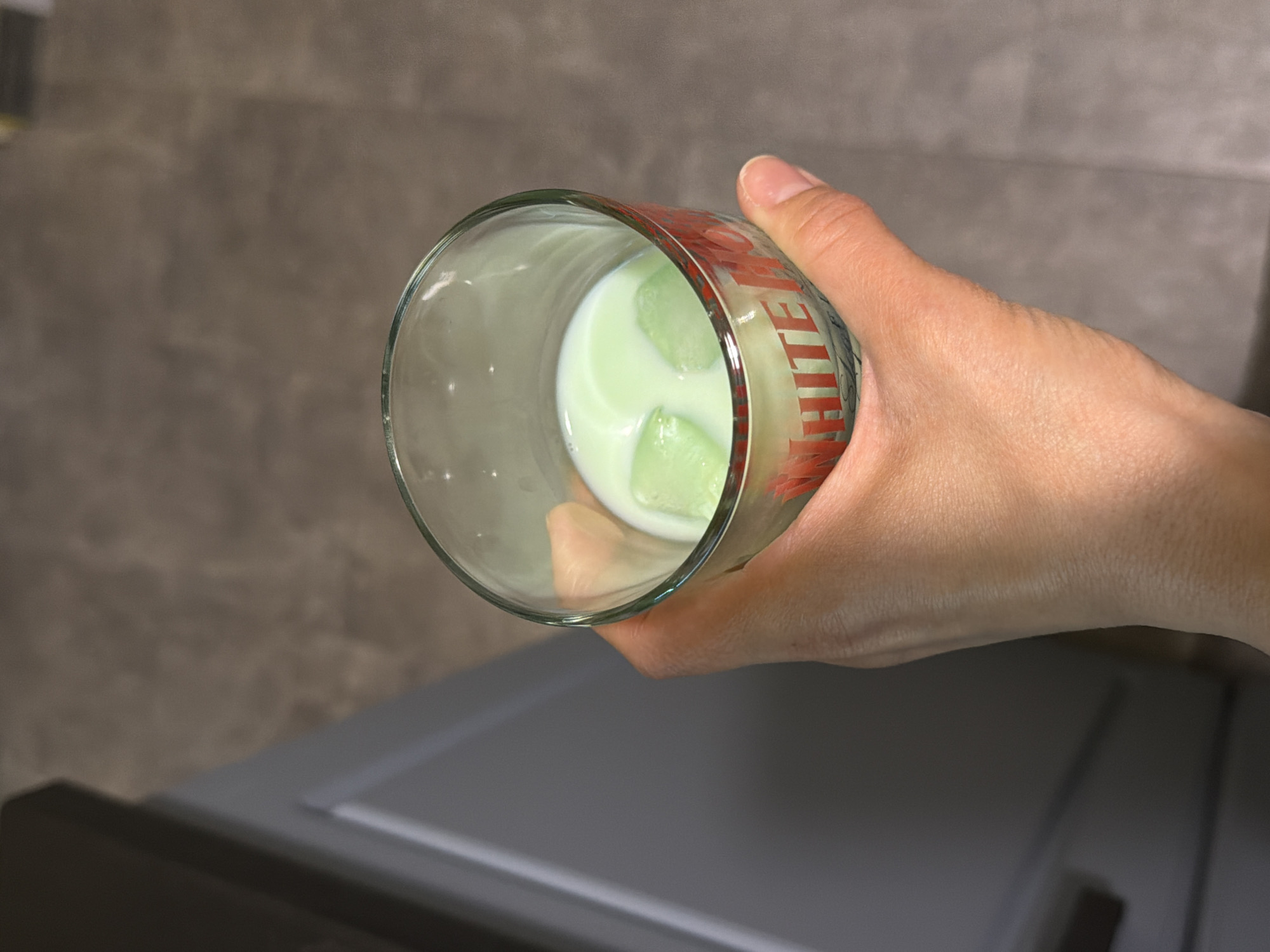 A person holds a glass of a pale green liquid with three ice cubes inside. The glass has red and white lettering, part of which reads "WHITE." The background is a grey wall and a grey surface. The city of Pleven, Bulgaria, is not discernable from this image.