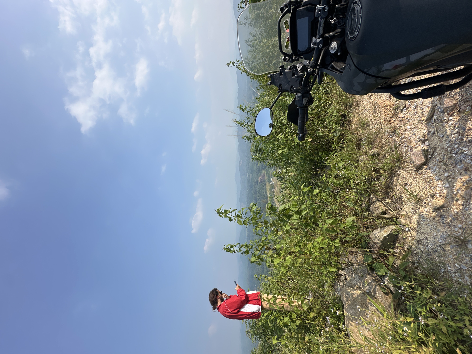 This landscape-oriented image captures a scenic outdoor moment, likely on a hill or mountain overlooking a valley in Tân Trịnh Commune, Vietnam.
In the midground, a person with dark hair, wearing a red and white jacket over light-colored pants, stands with their back mostly to the viewer, gazing down at a dark smartphone held in both hands. To their right, occupying the right side of the frame, is the front section of a dark-colored adventure motorcycle. Its clear windscreen, handlebars with controls, a side mirror (reflecting blue sky and greenery), and part of the fuel tank are visible.
The immediate ground around the person and motorcycle consists of gravel, dirt, and scattered rocks, interspersed with dense green vegetation, including shrubs and taller grass. Some small white flowers can be seen in the foreground. Beyond this, the background stretches into a vast expanse of light blue sky with scattered white clouds, and distant, hazy green hills or mountains, indicative of a natural, elevated landscape.
The scene takes place on a clear, sunny day, likely late morning or early afternoon, given the bright illumination and distinct shadows. The person appears to be taking a break from a motorcycle journey, possibly checking directions, capturing a photo, or communicating. The rugged motorcycle and the mountainous backdrop are strong location cues. No clear text is visible anywhere in the image.