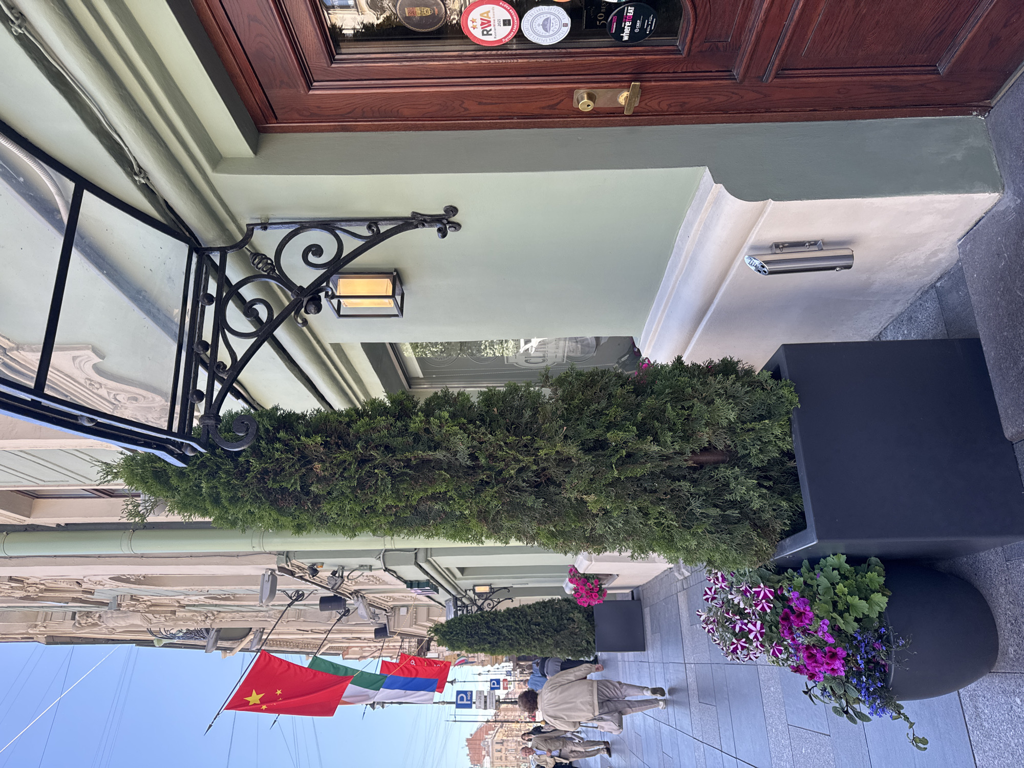 The image shows a storefront in Saint Petersburg, Russia, with flags of China and Russia flying from the building. The storefront features a dark wooden door with several circular stickers, including one that reads "where to eat." A glass awning with ornate black metal supports extends over the entrance. Two tall evergreen trees in dark planters flank the entrance. To the left of the entrance, a sidewalk is visible with several people walking and potted flowers. The building's facade is light green with decorative architectural details. The sky is clear and blue.