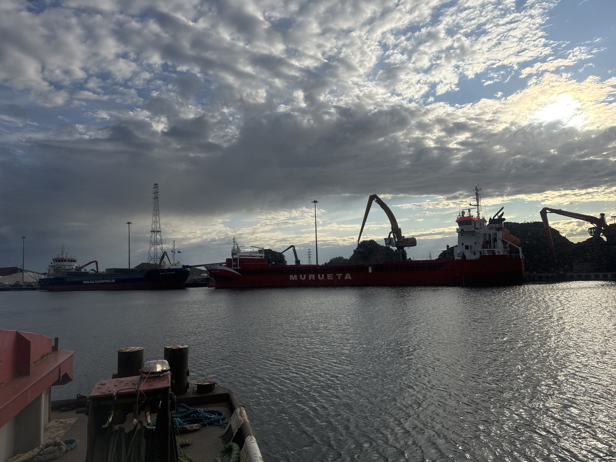 This image depicts an industrial port scene, likely within the city of Gdańsk, Poland. In the foreground, the stern of a dark-colored vessel is visible, featuring prominent mooring bollards and a small winch with coiled rope. The water surface is calm with gentle ripples, reflecting the overcast sky. In the mid-ground, a long, red cargo ship named "MURUETA" is docked. This vessel appears to be involved in the loading or unloading of bulk materials, as indicated by two large industrial cranes positioned alongside it, actively handling piles of scrap metal or similar debris on the shore. To the left of the "MURUETA", another vessel, a darker cargo ship named "BALTIC ECLIPSE", is also moored. The background is dominated by a dramatic sky filled with a mix of dark, heavy clouds and brighter, sunlit patches. Several tall industrial towers, likely power transmission pylons, are visible on the horizon. The overall atmosphere is one of active industrial work within a port environment under a dynamic sky.