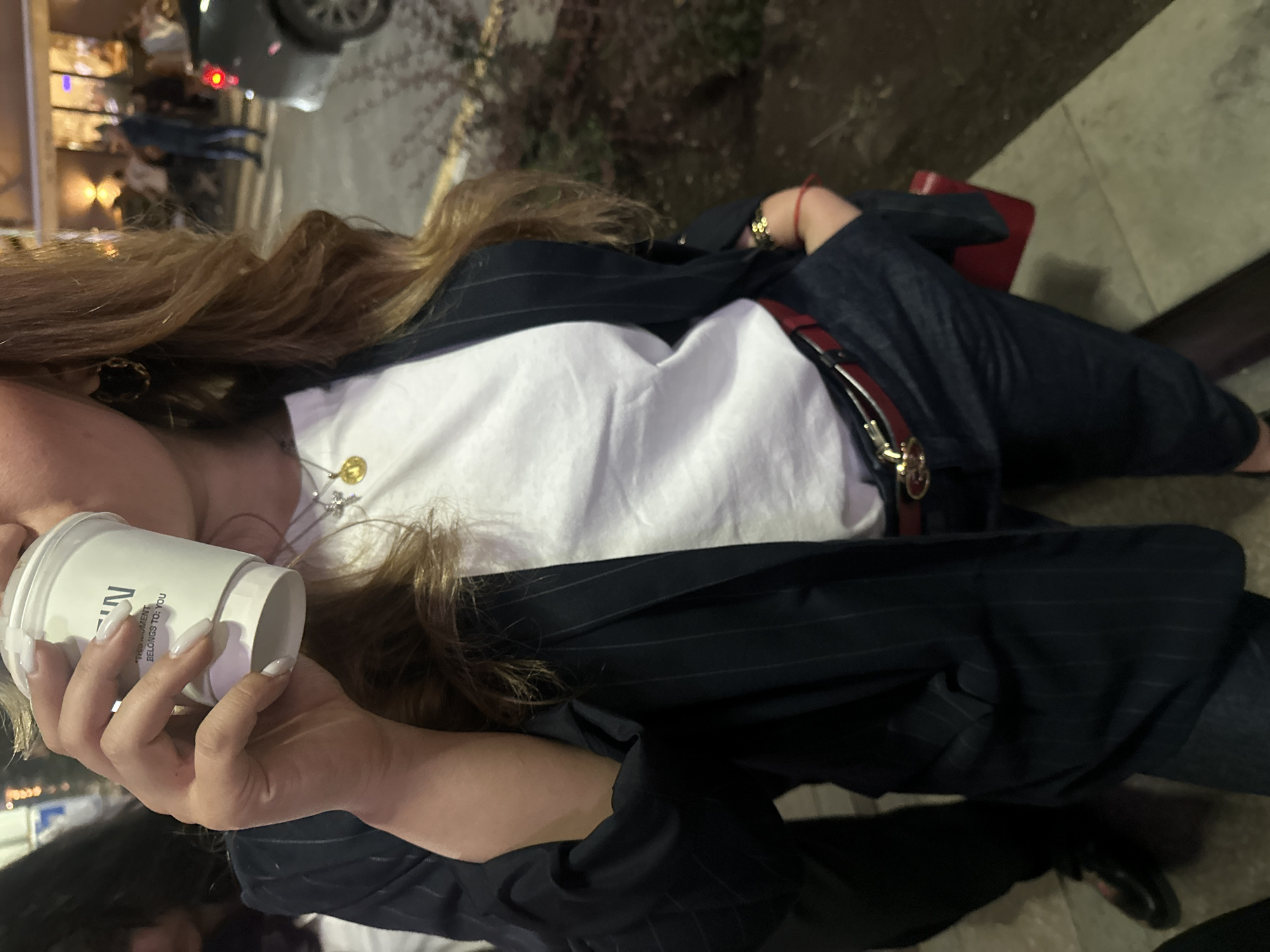 A person stands on an outdoor surface, holding a white cup in their left hand. They are wearing a white shirt under a pinstriped blazer, and dark denim pants held up by a red belt with a gold buckle. The person has long, light brown hair. In the background, blurred lights suggest a city street setting. The location is Yerevan, Armenia.