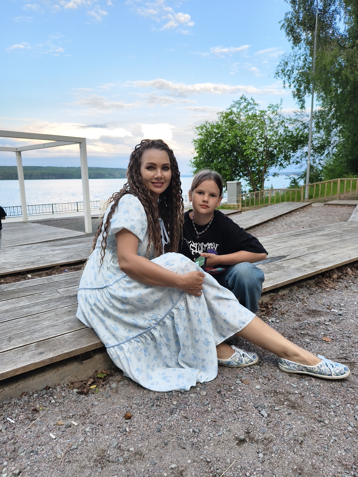In Chaikovsky, Russia, two individuals are seated on wooden planks outdoors by a body of water. The person on the left is an adult female with long, curly brown hair, wearing a light blue dress with a floral pattern and matching shoes. She is smiling and looking towards the camera. To her right sits a younger individual, likely a child or adolescent, with medium-length brown hair, wearing a black t-shirt and blue jeans. This person is looking towards the camera with a neutral expression and is holding a mobile phone. They are positioned on a wooden deck that appears to be near a waterfront promenade, with trees and a lake visible in the background under a cloudy sky.