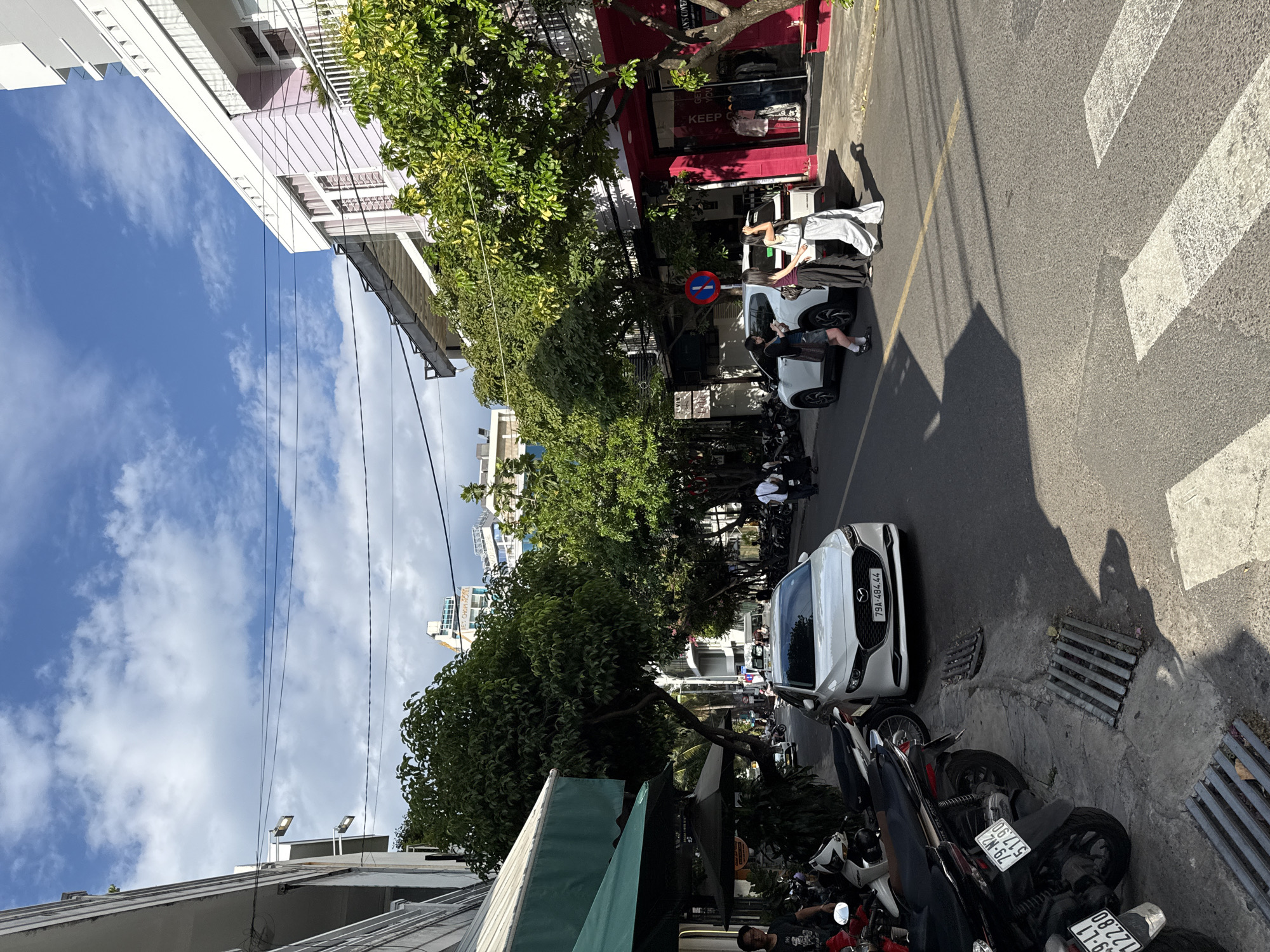 The image depicts a street scene in Nha Trang, Vietnam, on a sunny day with scattered clouds. The street is lined with buildings, some of which are brightly colored, including a pink and white building with balconies. Dense green trees shade the sidewalk. Several motorcycles are parked along the left side of the street, and a white car is parked on the right. Three young women are standing near the back of the white car, one of whom is looking at her phone. Another car is visible further down the street. Power lines crisscross above the scene. A crosswalk is marked on the asphalt in the foreground.