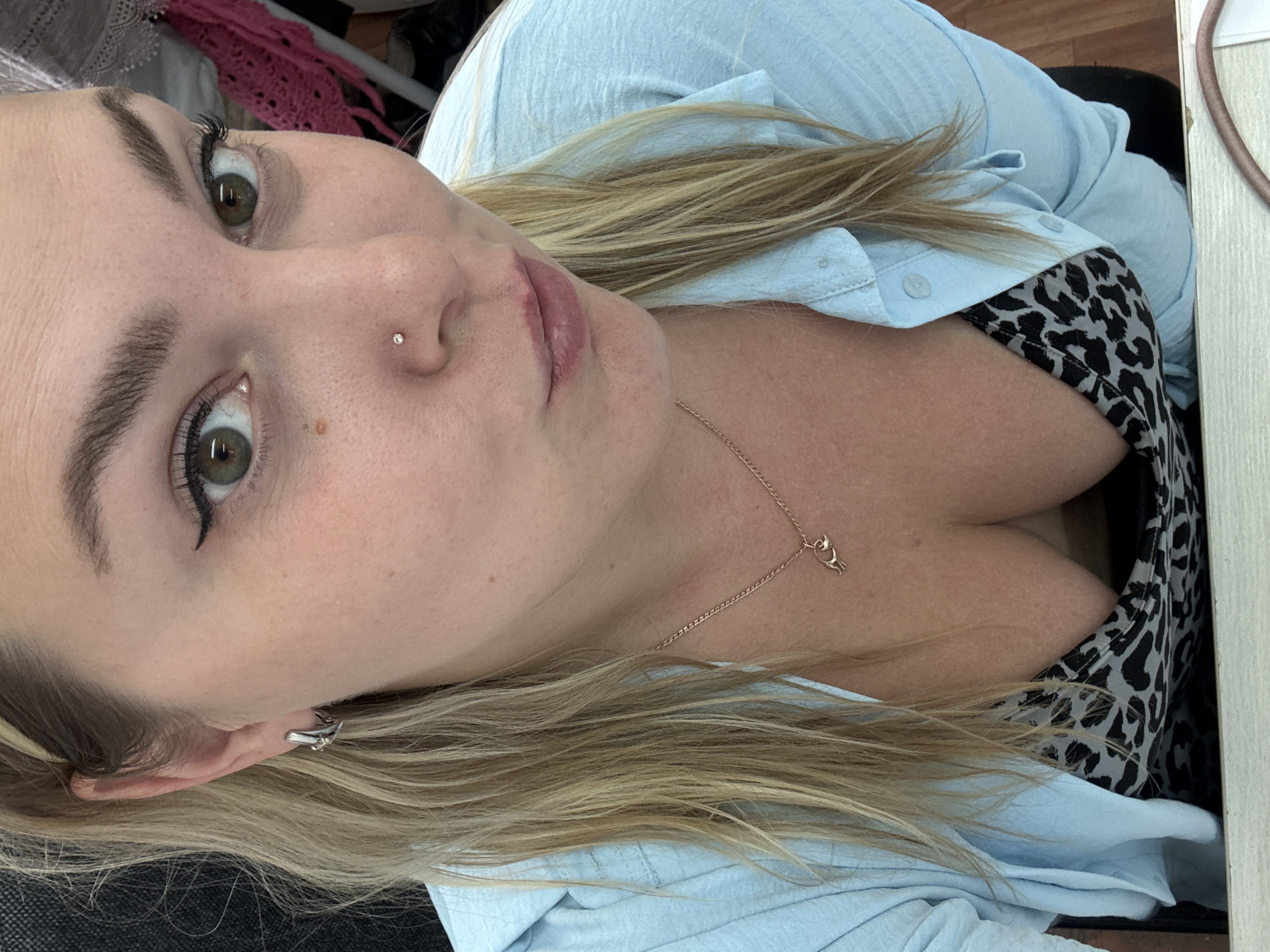A blonde woman with green eyes and a nose piercing is looking directly at the camera. She is wearing a light blue button-up shirt, partially open to reveal a leopard-print bra. A thin rose gold necklace with a small pendant is visible around her neck. Her hair is blonde and falls past her shoulders. The background is blurred, but appears to be an indoor setting with some fabric objects in pink and white. The image is a close-up, upper body shot of the woman, taken from a slightly low angle. There is no visible indication of the city of Noviy, Russia in the image.