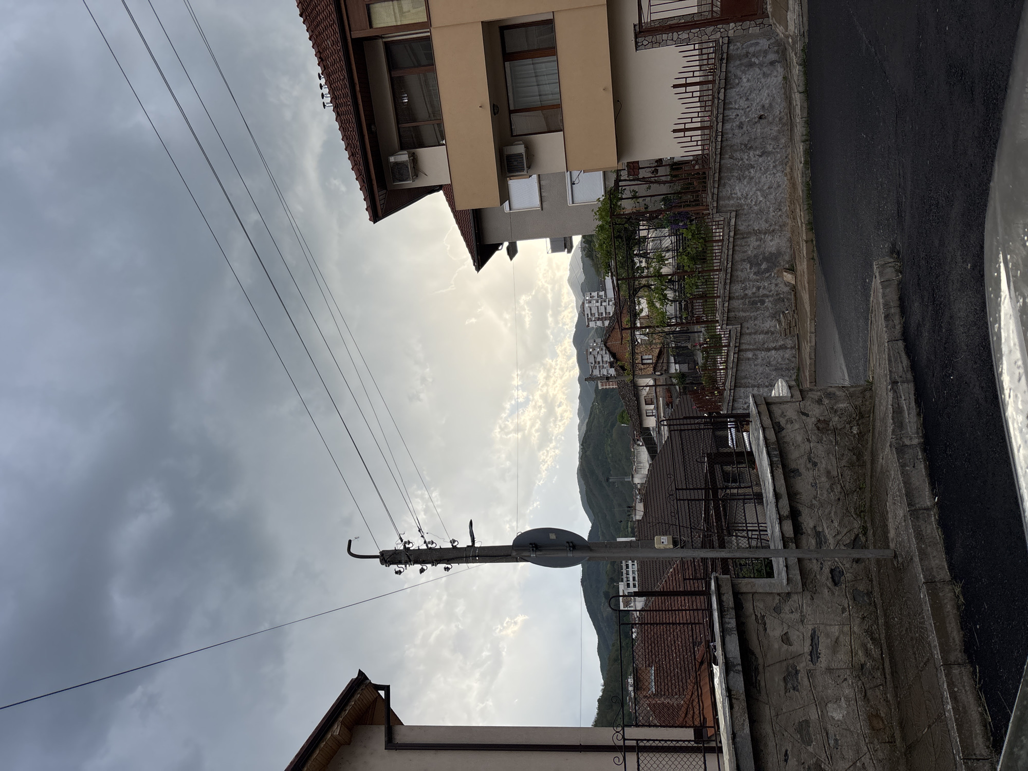 The image depicts a street scene in Zlatograd, Bulgaria. The viewpoint is from a slightly elevated position, looking down a narrow paved street. The weather appears overcast with a cloudy sky. Several buildings are visible, including a tan-colored building with multiple windows and air conditioning units, and a smaller building with a visible roof and a light-colored facade. A stone wall with a metal fence runs along the street. A utility pole with electrical wires and a sign is in the foreground. Mountains and other buildings are visible in the distance.