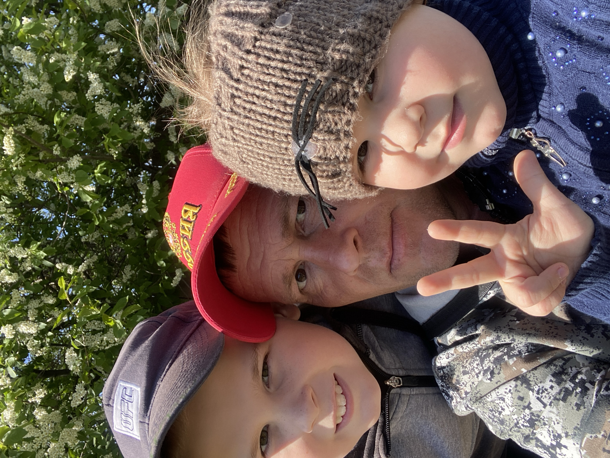 The image shows a close-up selfie of a man with two children. The man is wearing a red baseball cap, and he is positioned in the center of the frame. One child is wearing a gray knitted hat with cat ear details, positioned above the man, while the other child wears a gray baseball cap and is positioned below. The man is making a peace sign with his hand, and the children are smiling. The background is filled with green foliage and white flowers, suggesting they are outdoors. The image doesn't have enough information to definitively confirm the location is Фоки, Russia. However, if the location information provided is accurate, the subjects are most likely located outdoors in a natural environment, possibly a park or garden, given the surrounding foliage.