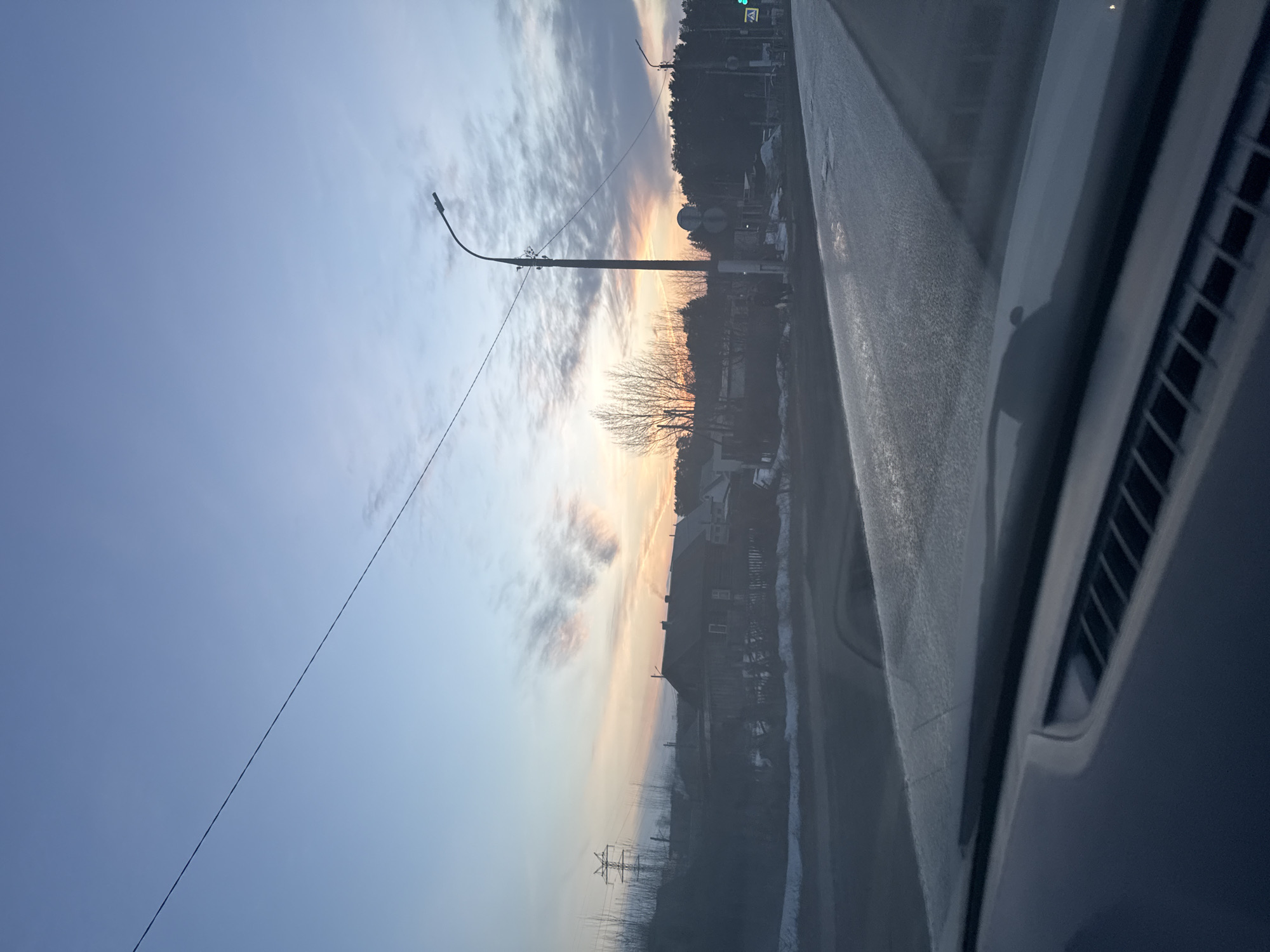 The image, taken from inside a vehicle, shows a street scene in Chaikovsky, Russia at dusk. A sky transitions from blue to orange. A utility pole stands along the street. There are trees and buildings visible. The image is somewhat blurry due to the perspective through a car window.