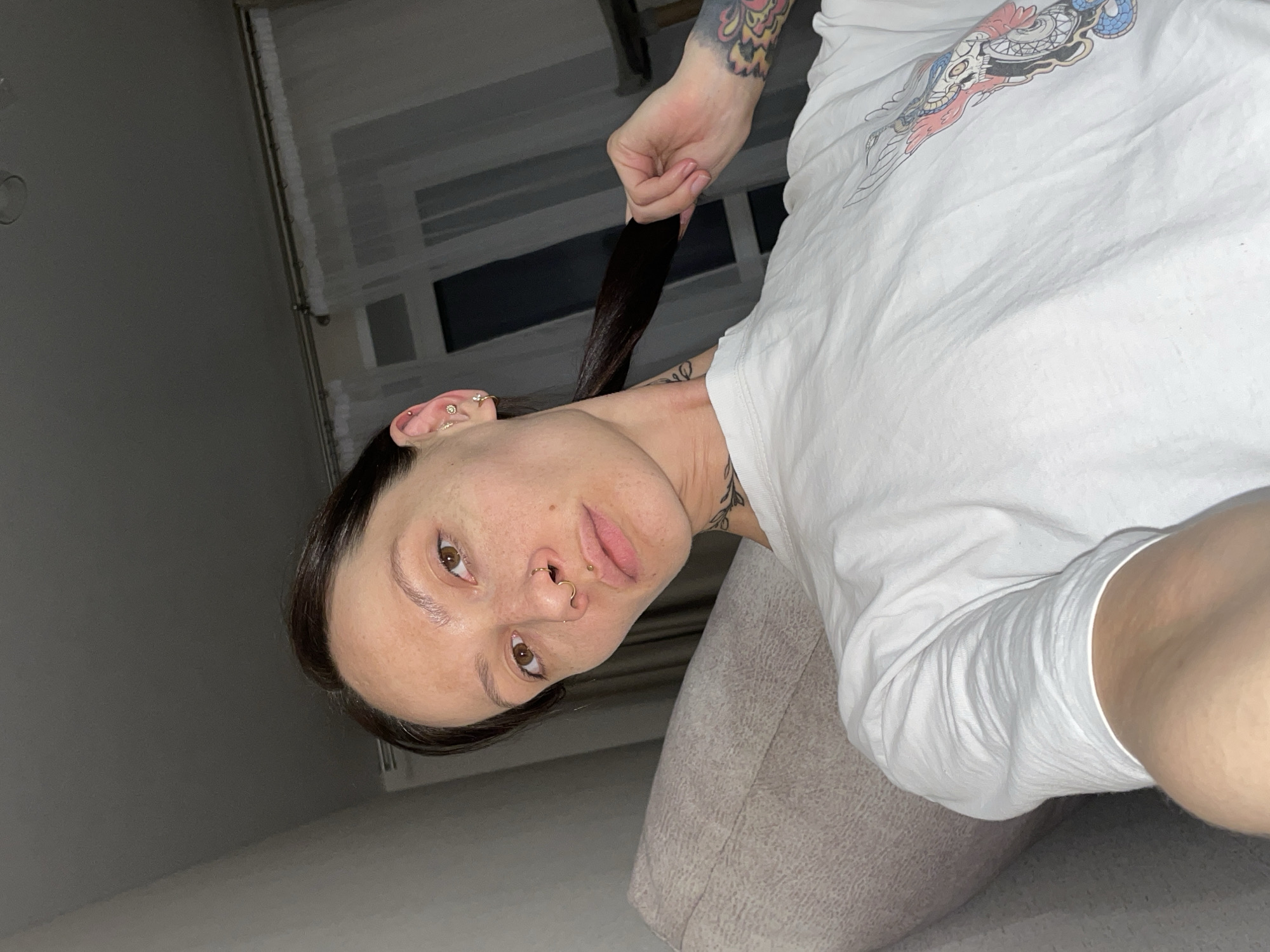 A woman with dark hair pulled into a ponytail is holding the end of her ponytail with her right hand. She is wearing a white T-shirt with a graphic design on the upper right side. She has multiple piercings in her left ear and nose rings. Her gaze is directed towards the camera. She is sitting on a beige couch in what appears to be an indoor setting. There is a window in the background, and a wall is visible on the left side of the image. The location in the city of Izhevsk, Russia, is not discernible from this image.