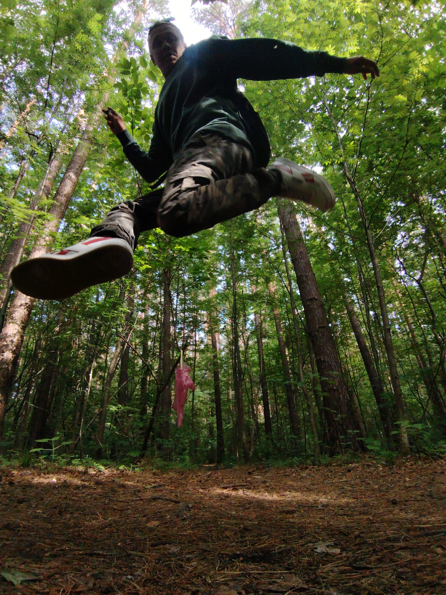 A dynamic photograph, taken from a low angle, captures a person mid-jump in a dense forest in Chaikovsky, Russia. The individual, appearing male, is silhouetted against the bright, sunlit sky peeking through the canopy of tall trees. He wears a dark long-sleeved shirt and camouflage cargo pants, with white sneakers featuring red details clearly visible on his feet. His arms are outstretched, and legs are bent, emphasizing the apex of his leap.
The setting is a vibrant forest, likely deciduous or mixed, with numerous tall tree trunks rising from the forest floor. The ground is covered with fallen leaves, pine needles, and sparse undergrowth, suggesting a natural, undisturbed environment. Sunlight filters through the leaves, creating a dappled effect on the forest floor. A notable detail in the mid-ground is a bright pink or red plastic bag or piece of fabric hanging from a tree branch, providing a pop of contrasting color against the greens and browns of the woods. The time of day appears to be bright daytime, likely sunny. There is no visible text in the image.