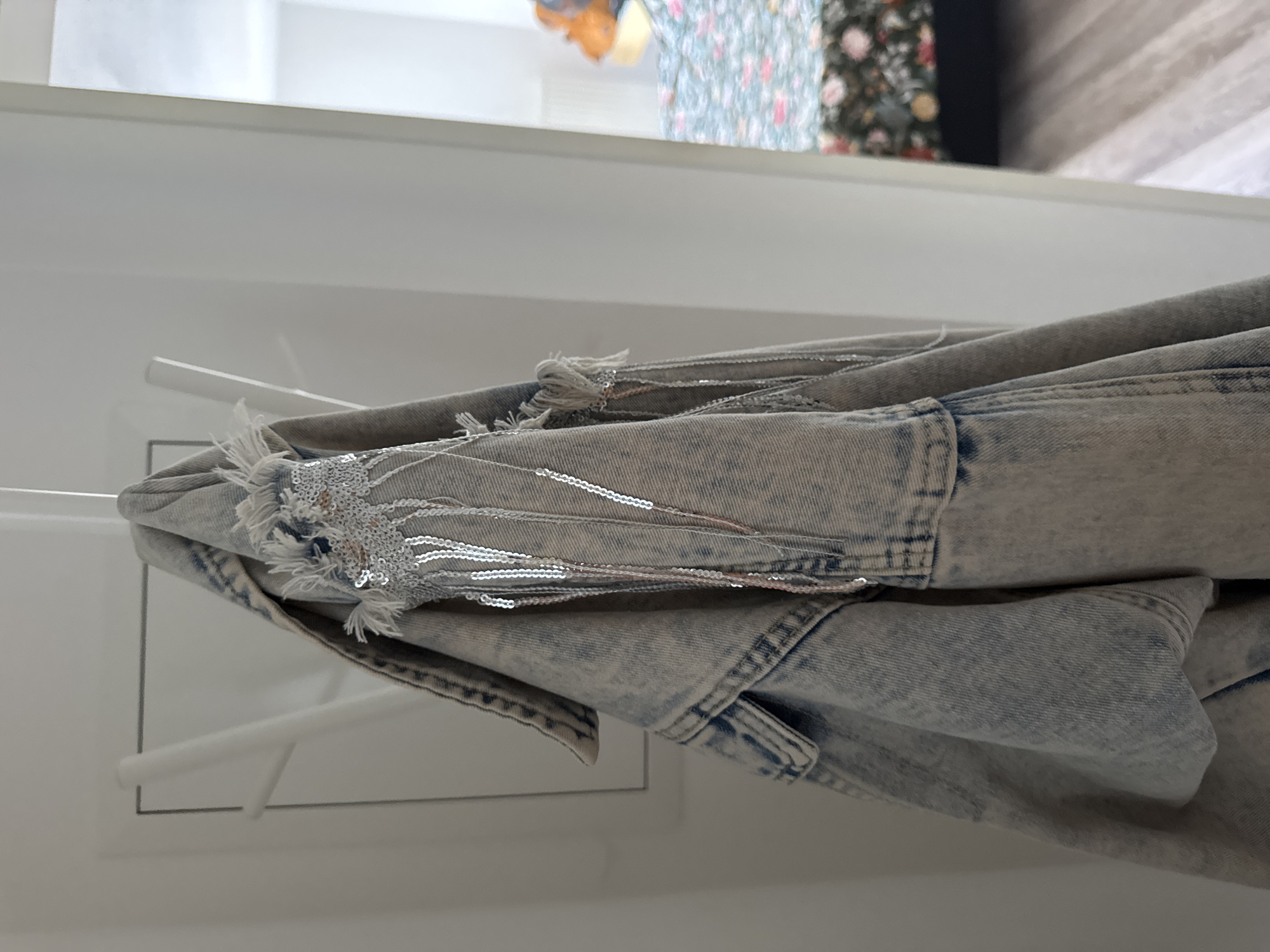 The image shows a close-up of a light-wash denim jacket with distressed detailing and dangling silver sequins, hanging on a white coat rack. The jacket is positioned on the left side of the frame, with the right side of the image showing a slightly out-of-focus view of a bedroom with a floral-patterned bedspread. The lighting is soft and natural. The scene is located in a room in Höhenkirchen-Siegertsbrunn, Germany.