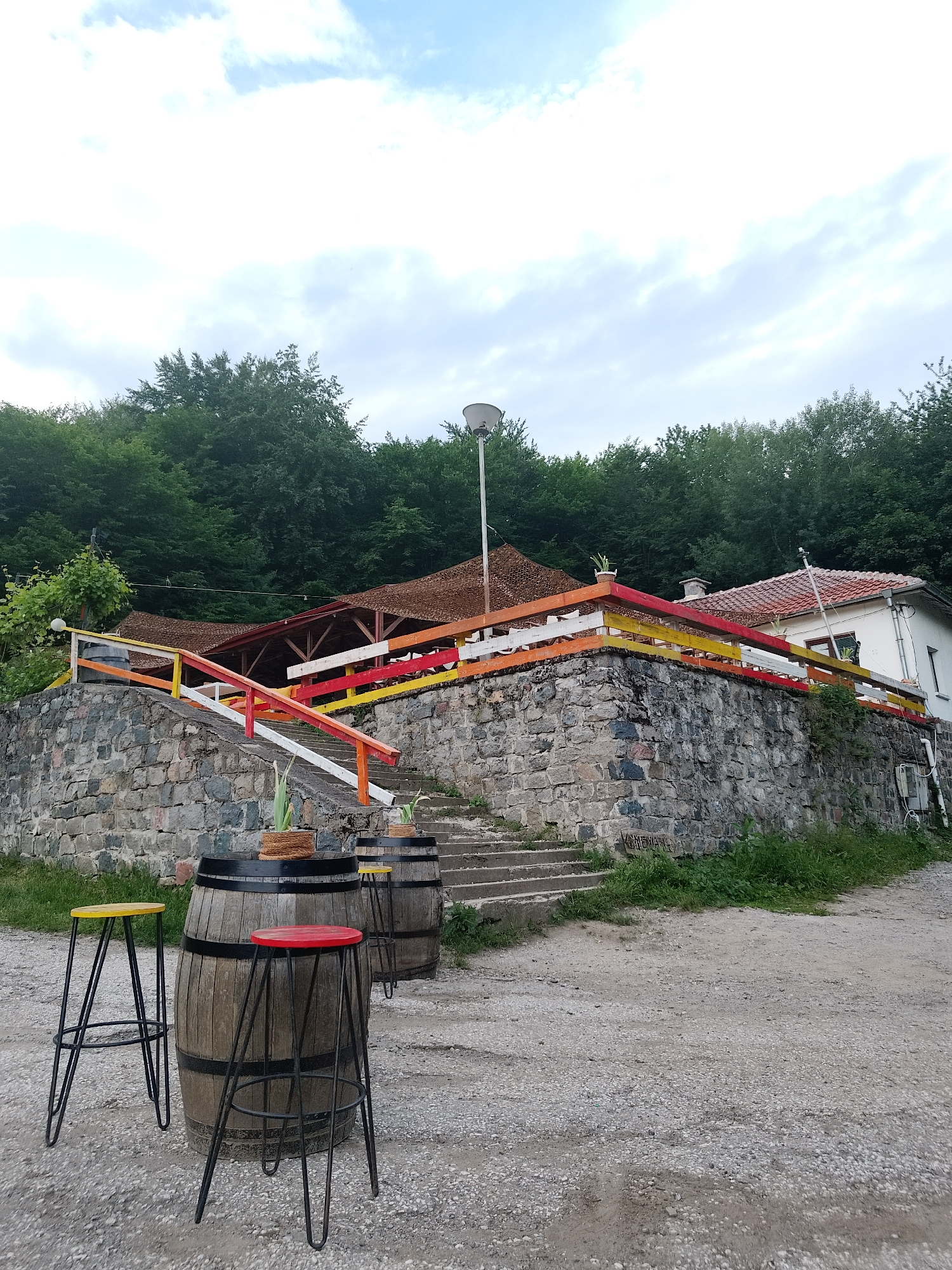 The image shows an outdoor seating area with two wooden barrels and three metal bar stools in the foreground. The barrels appear to be used as tables, with one having two potted plants on top. The bar stools have red and yellow seats. Behind the barrels and stools, a stone wall with a colorful railing runs along a set of outdoor stairs. The railing is painted in alternating sections of yellow, orange, red, and white. Further up the stairs, a building with a red tile roof is visible, and behind that, a dense forest covers a hillside. A lamppost stands near the building. The ground is a gravelly surface. The scene is set under a cloudy sky. This appears to be a rustic establishment, possibly a restaurant or bar, in Sofia, Bulgaria.