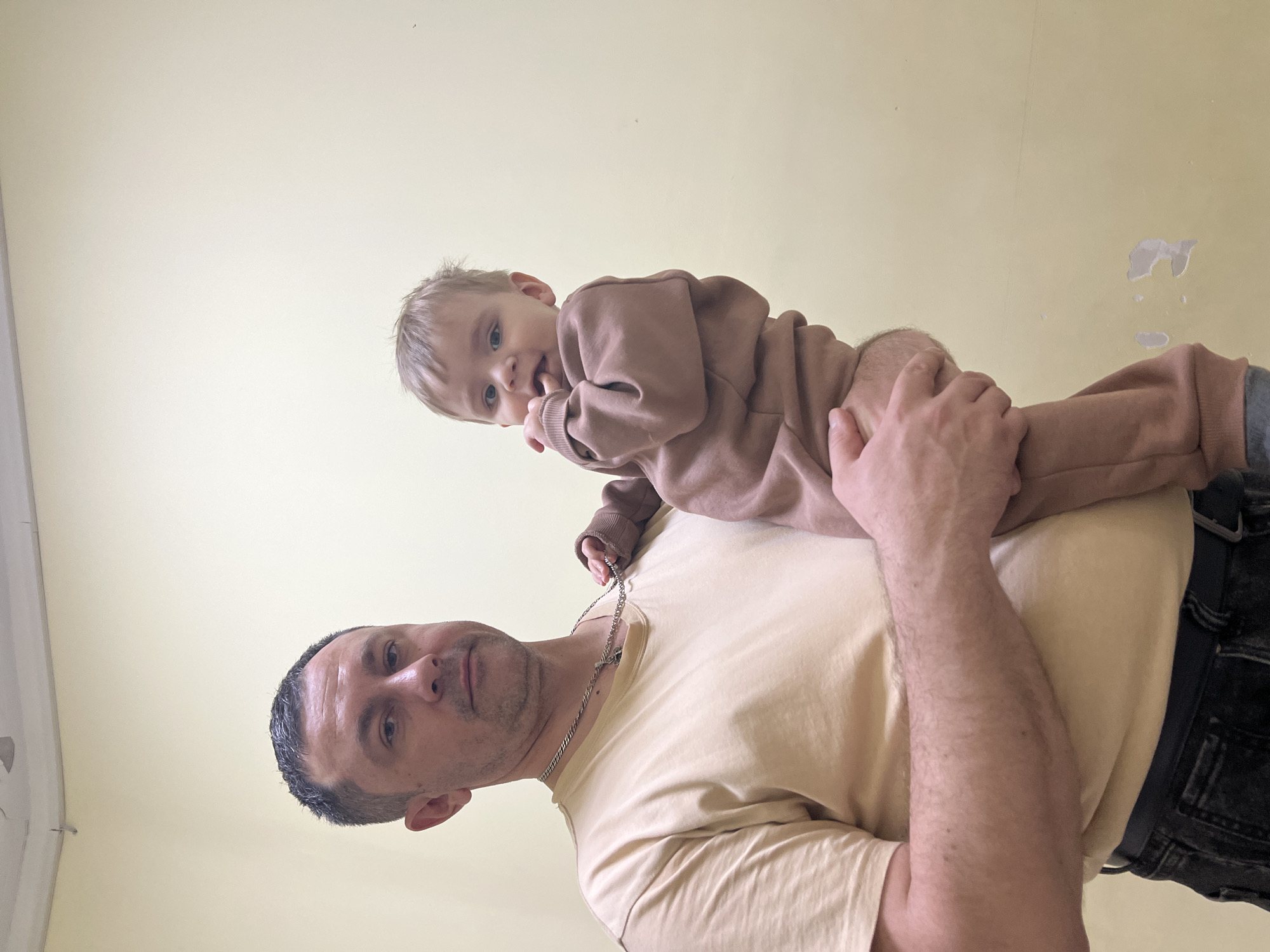 A man is holding a young child, approximately 1-2 years old, on his shoulders. The man is fair-skinned with short, graying hair and is wearing a light beige t-shirt and dark jeans with a belt. He has a silver chain around his neck. The child is wearing a light brown, long-sleeved outfit and has blonde hair. The background is a pale yellow wall with some minor imperfections. The location is unspecified and there is no reference to Noviy, Russia.