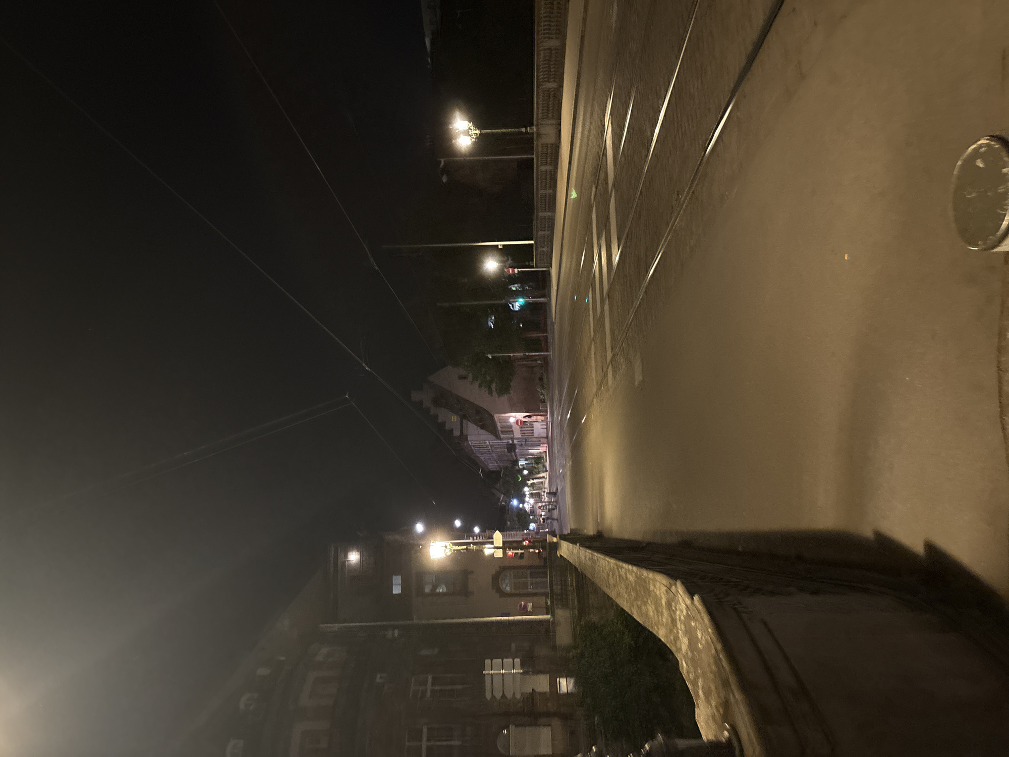 The image depicts a night scene on a bridge in Strasbourg, France. The view is oriented along the bridge's length, with the road surface dominating the right side of the frame. Parallel tram tracks run along the road. A low wall lines the bridge's edge.
Across the road, buildings are visible on the left, illuminated by streetlights. The streetlights cast light onto the road and sidewalks, suggesting a residential or commercial area.
In the distance, streetlights line the route, indicating continued urban development. People can be seen as blurred figures in the background.
The sky is dark, with utility wires crossing the frame.