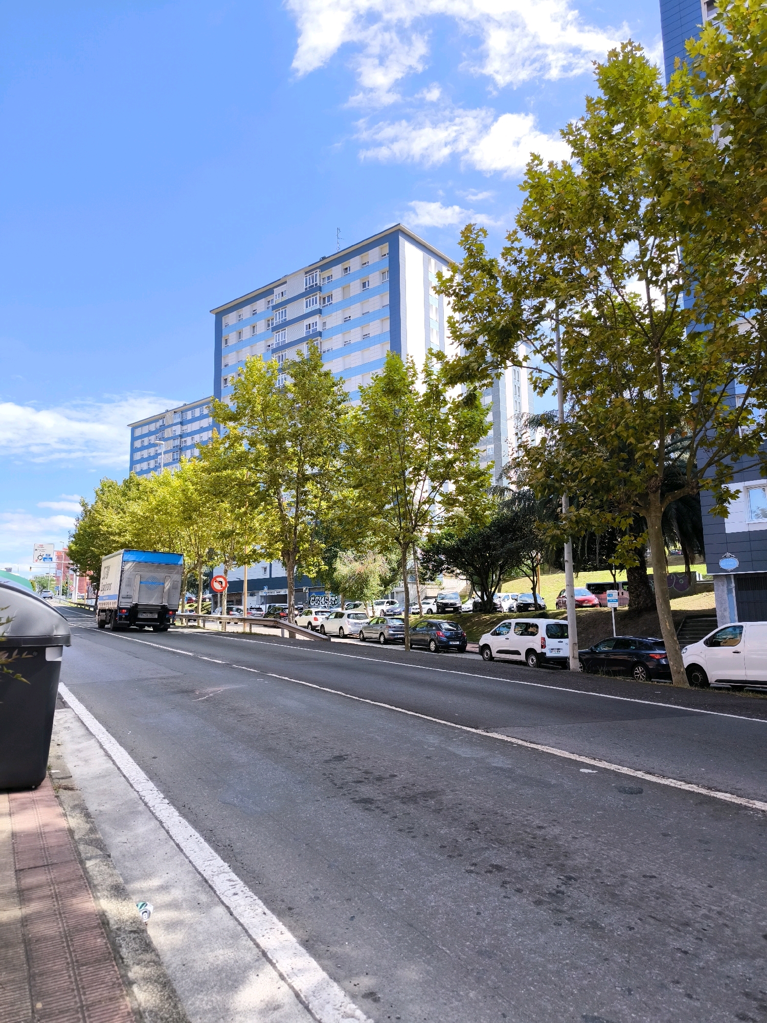 The image captures a street scene in Barakaldo, Spain, on a bright, sunny day. A multi-lane asphalt road dominates the foreground, with a sidewalk lined with red brick paving and a gray trash bin on the left. A white line delineates the edge of the road. Parked cars are visible along the right side of the street, including a white van, a dark car, and several other vehicles. Across the road, a row of trees with green foliage shades the sidewalk and the adjacent buildings. These trees are positioned in front of a modern, multi-story building complex with a distinctive blue and white facade. Further down the street, a white truck with a blue stripe is driving away from the camera. The sky is a vibrant blue, dotted with scattered white clouds. The scene appears to be a residential or commercial area within the city.