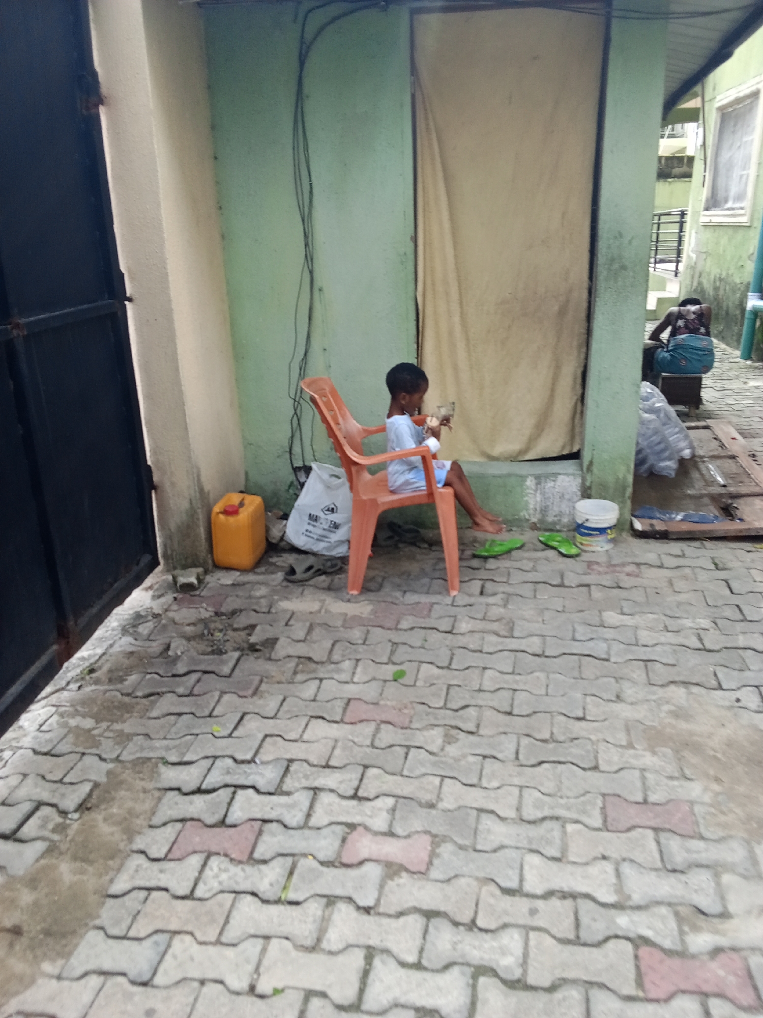 This image depicts an outdoor residential scene in Ado, Nigeria, characterized by a paved ground made of interlocking concrete blocks, predominantly gray with intermittent reddish-brown sections. On the far left, a dark metal gate or door is partially visible next to a beige-painted wall showing dark staining at its base. Adjacent to this wall, a bright yellow plastic jerrycan rests on the ground.
In the center, a young child, approximately 4-7 years old, with dark hair and wearing a light blue t-shirt and shorts, is seated barefoot on an orange plastic chair. The child holds a small, light-colored object near their face, appearing to interact with it. The chair is positioned against a light green wall from which dark electrical wires descend vertically. Behind the child, a large, light-colored fabric or curtain hangs in a doorway opening within the light green wall. On the ground directly in front of the child are two bright green flip-flops and a white plastic bucket with a lid. To the right of the bucket, a translucent bundle of items, possibly plastic bottles, is visible, along with a wooden board.
Further in the background on the right, an adult figure is seen from behind, seated on a dark stool or block. This individual wears a patterned top and a blue lower garment. Behind this person, another light green building features a white-framed window and a dark metal railing. The overall environment appears functional and lived-in, with visible wear on surfaces.