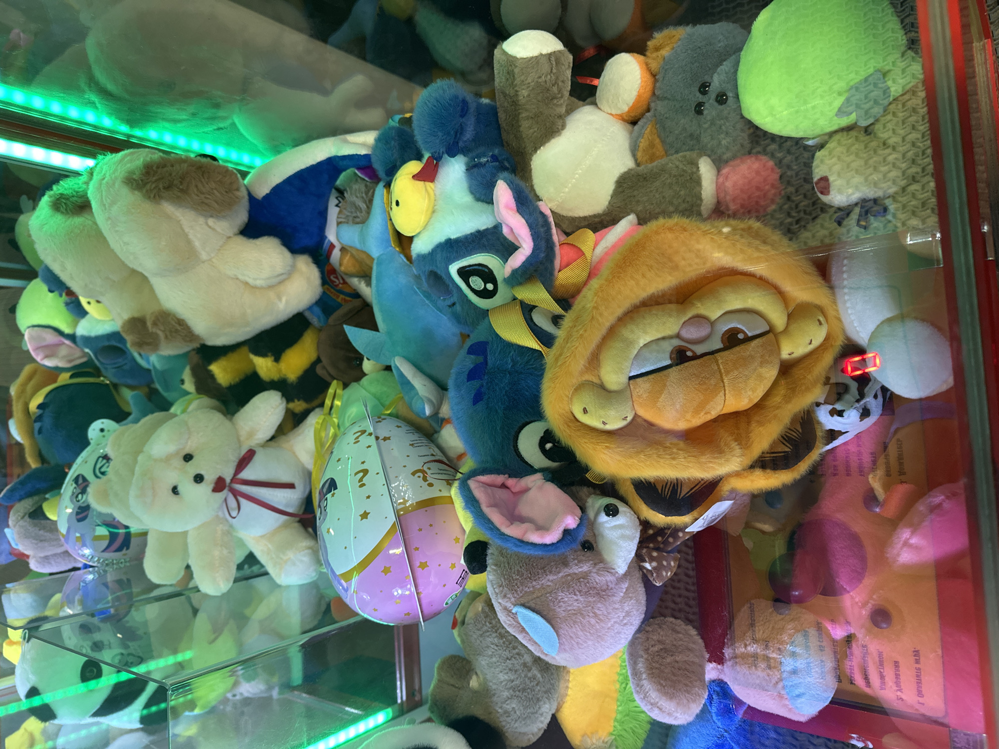 The image shows a collection of stuffed toys inside a claw machine, likely in an entertainment venue or arcade. The toys are varied, including a panda, bears, a shark, a duck, and a character resembling Garfield. The lighting is bright, with green neon lights visible on the right side of the machine. The toys are densely packed, with some appearing to be partially obscured by others. The machine has a glass front through which the toys are visible. The interior of the machine shows a colorful and chaotic arrangement of plush toys, typical of a prize vending machine. There are no people visible in the image, nor any specific indicators of the exact location within Minsk, Belarus beyond it being an indoor establishment.