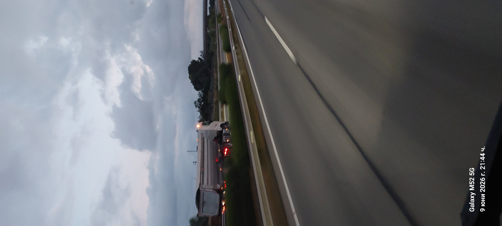 A white dump truck is traveling on a highway in Vila-seca, Spain. The truck is carrying a load of dark material and has its headlights and brake lights on. The highway is paved and has white lines marking the lanes. There are trees and vegetation on either side of the highway. The sky is cloudy and appears to be dusk or early evening. The text overlay indicates the image was taken on June 9, 2026, at 21:44.