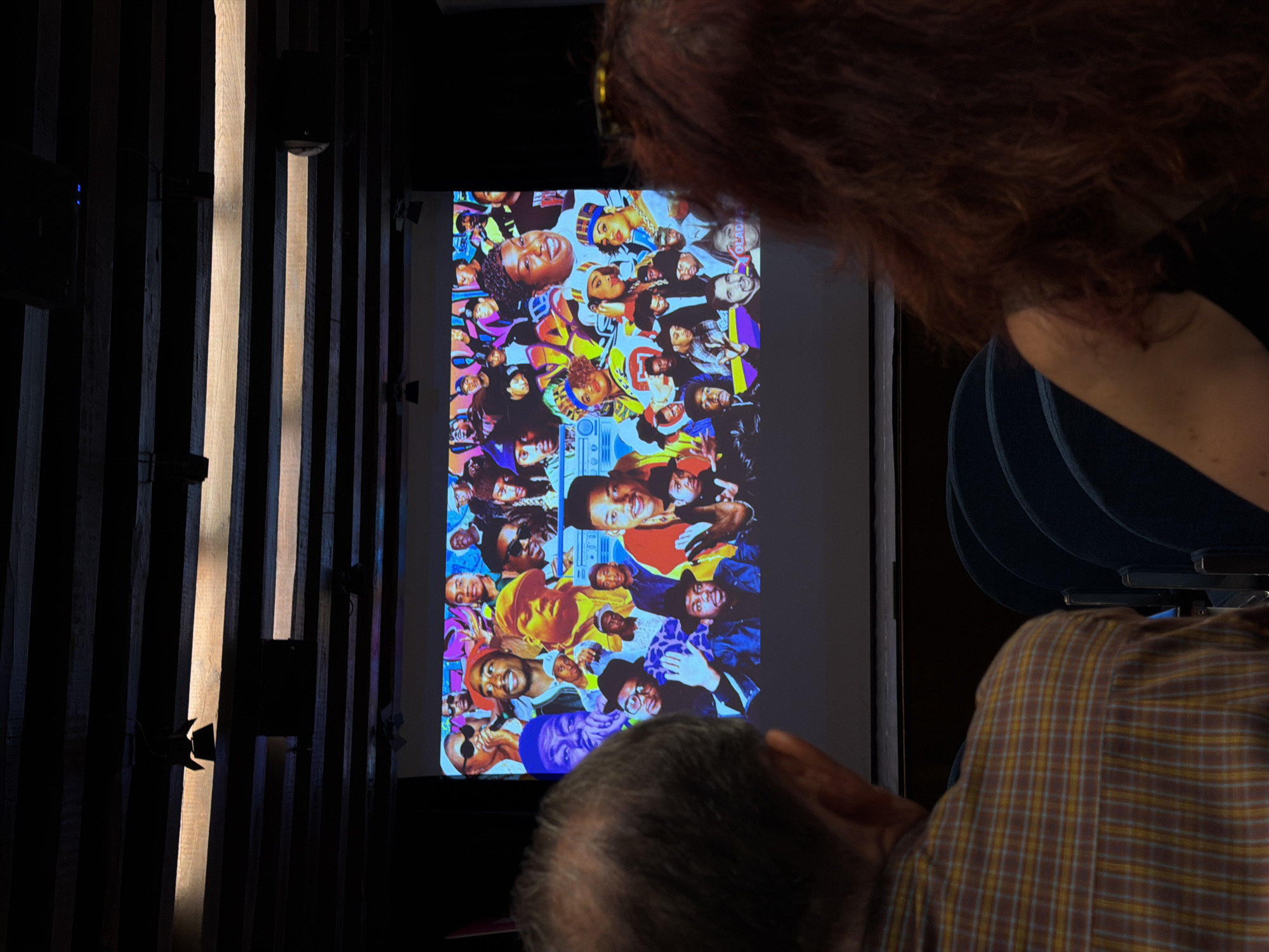 In San Lorenzo de El Escorial, Spain, two individuals, viewed from behind, observe a projected image on a large screen. The projection displays a colorful collage of faces, primarily featuring prominent figures from the hip-hop community. The audience is seated in a dimly lit room, suggested by the dark ceiling with wooden beams and lighting fixtures. The man on the left wears a plaid shirt, while the woman on the right has long, reddish-brown hair. The scene appears to be an indoor event, possibly a presentation or screening, taking place in a venue with tiered seating visible in the foreground.