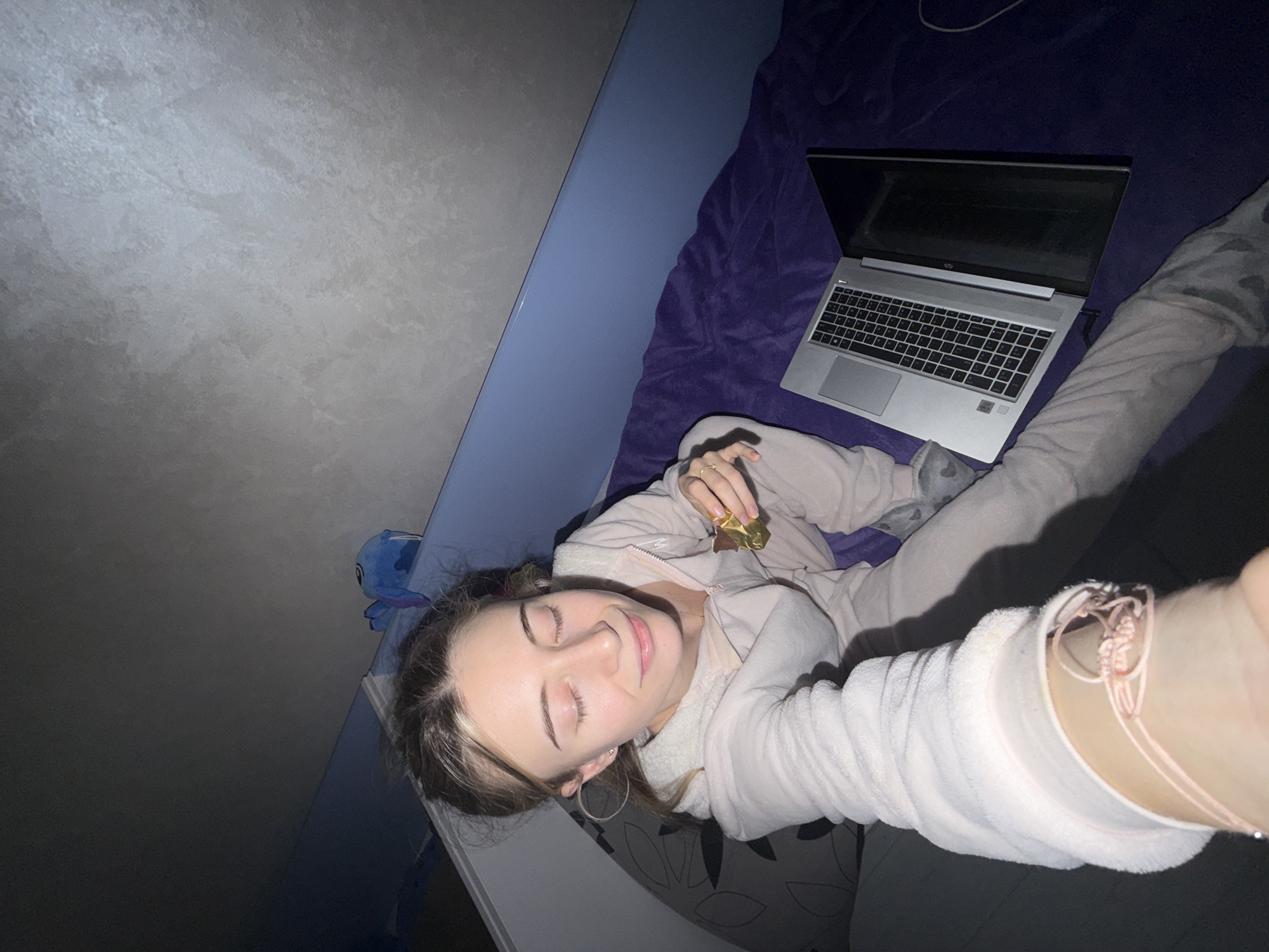 A young woman is lying on a bed in what appears to be a bedroom. She is wearing a light pink zip-up hoodie and matching pants. She is holding a wrapped chocolate in her right hand. A silver laptop is positioned on the bed near her. The bed has a purple duvet. The wall is a light grey color with a blue bed frame. There is a Stitch stuffed animal visible in the background. The image does not provide any information indicating this scene is in Plovdiv, Bulgaria.