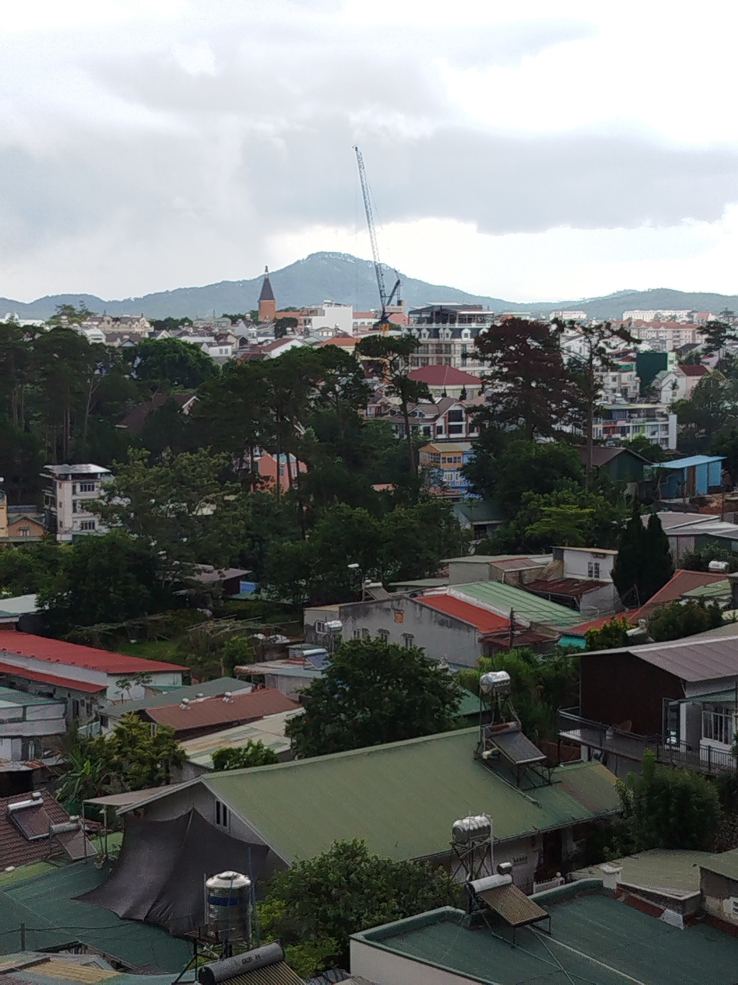 The image presents an elevated view of a section of Da Lat, Vietnam. Dominating the foreground are rooftops of houses and buildings, many with green roofing, and some sporting solar water heaters. Interspersed among the buildings are trees, lending a verdant quality to the scene. In the middle distance, buildings become more densely packed.
In the background, a church steeple is visible, along with a construction crane. Hills are silhouetted against an overcast sky, which appears predominantly gray. There are no people visible in the image. The time of day is daytime, based on the ambient lighting.