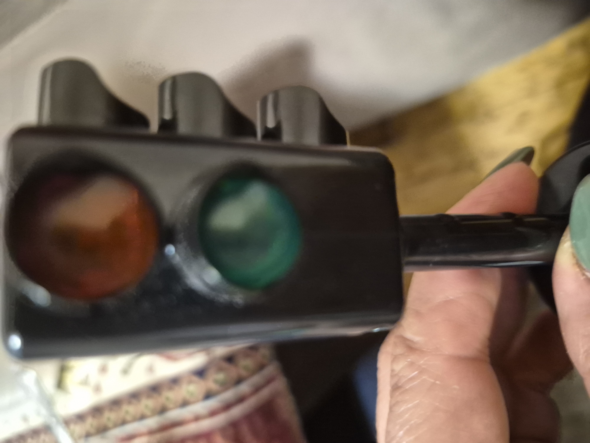The image displays a close-up, slightly blurred shot of a person's right hand holding a small, black, handheld device resembling a traffic light. The device features two circular lights on its front: a red or orange light on the left and a green light on the right, both appearing illuminated. On the top edge of the device, there are three small, dark, knob-like elements. A black handle or stem extends from the right side, which is gripped by the fingers of the hand.
The person's fingernails are painted a distinct light green or teal color. The skin of the hand shows visible texture and creases. The background is out of focus, creating a shallow depth of field, with the traffic light and hand as the primary subjects. Partially visible in the lower left background is a blurred, patterned rug or textile. Above and behind the device, indistinct blurred surfaces are visible, including a light-colored area and a yellowish-brown area, likely indicating indoor furnishings. There is no visible text in the image. The scene is indoors, and no specific time of day or discernible emotions are indicated. The location context provided is Yerevan, Armenia, though this is not visually identifiable from the image content.