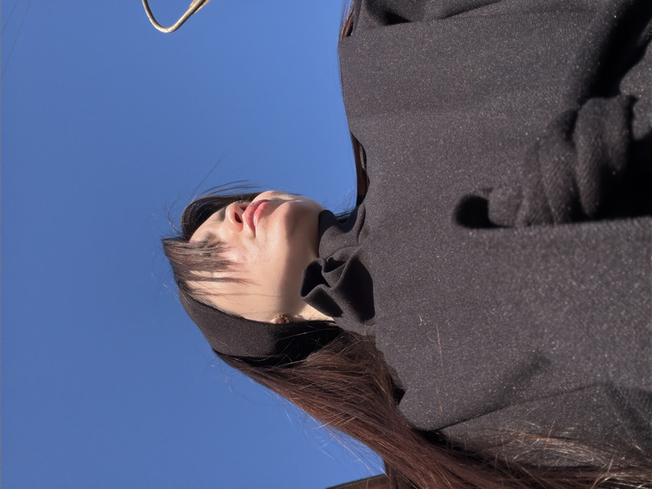 A single individual is depicted in a low-angle outdoor shot against a clear blue sky, in the city of Chaikovsky, Russia. The person, appearing female, has long, dark hair, partially covered by a black headband. They are wearing a dark, textured garment with a high, gathered collar. A small, light-colored earring is visible on the right earlobe. The individual's head is tilted upwards and to the right, with direct sunlight illuminating the right side of their face, creating a shadow along the jawline. Their lips are slightly parted or pursed. An indistinct dark object is partially visible in the upper left background, and a curved, light brown object is present in the upper right background. The environment is brightly lit.