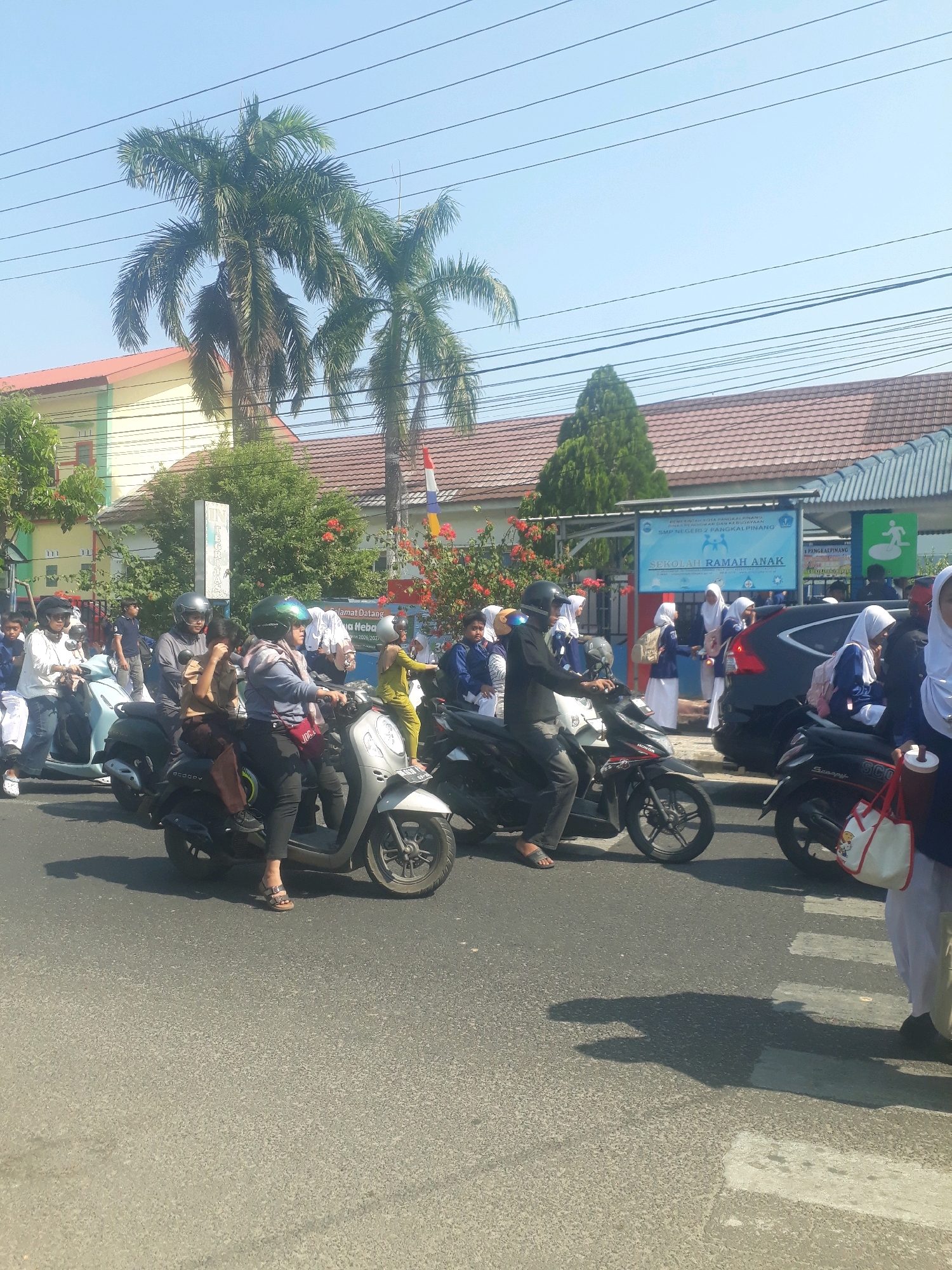 The image depicts a street scene in Pangkalpinang, Indonesia, likely near a school as indicated by the students on motorcycles and the school building in the background. The sky is clear and blue, with power lines crisscrossing overhead. Two palm trees are visible on the left side of the image, with lush green foliage.
A group of people, predominantly students wearing school uniforms, are seen on several motorcycles on a paved road. Some are wearing helmets for safety. One motorcycle in the foreground has two students, one driving and one seated behind. Another motorcycle to the right also carries two students. Other students are visible in the background, some walking or waiting near a building.
The building in the background appears to be a school, with a sign that reads "SEKOLAH RAMAH ANAK" (Child-Friendly School). There is also a black car parked near the school entrance. The environment is sunny and appears to be a typical day in the city. The road has white pedestrian crossing lines.