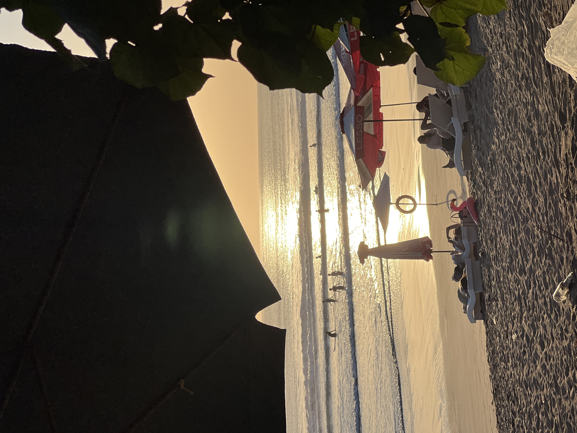 This image captures a serene sunset scene at Kuta Beach in Bali, Indonesia. The sun, low on the horizon, casts a warm, golden glow across the ocean and wet sand, creating a shimmering reflection. Several people are visible in the shallow waves, some with surfboards, enjoying the surf.
In the foreground, the beach is dotted with lounge chairs and umbrellas, offering a place for relaxation. Two individuals are seated on lounge chairs under a red umbrella, appearing relaxed. A pink flamingo float adds a playful touch to the scene. The sand itself appears well-trodden, with a few scattered items, including a bottle and some debris.
The image is framed by the dark silhouette of what appears to be a large umbrella or canopy in the upper left, and lush green foliage on the right, adding depth and a sense of enclosure to the beach setting. The overall atmosphere is tranquil and idyllic, characteristic of a beach sunset.