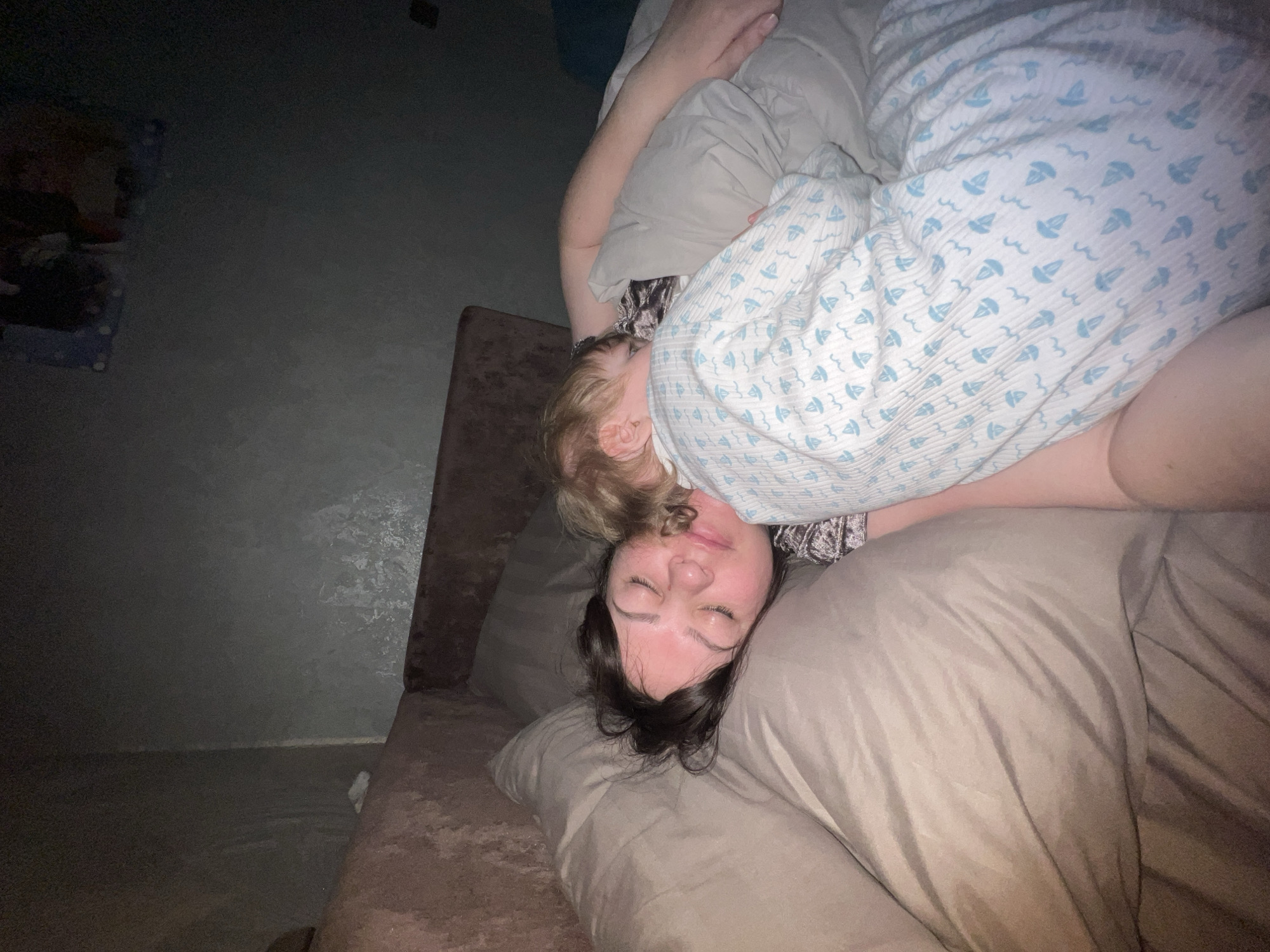 A photograph depicts an adult woman lying on a bed, with a small child lying on top of her. The woman is lying on a beige pillow with her eyes closed. The child is partially covered by a white blanket with a sailboat pattern. The location is Noviy, Russia is not discernible.