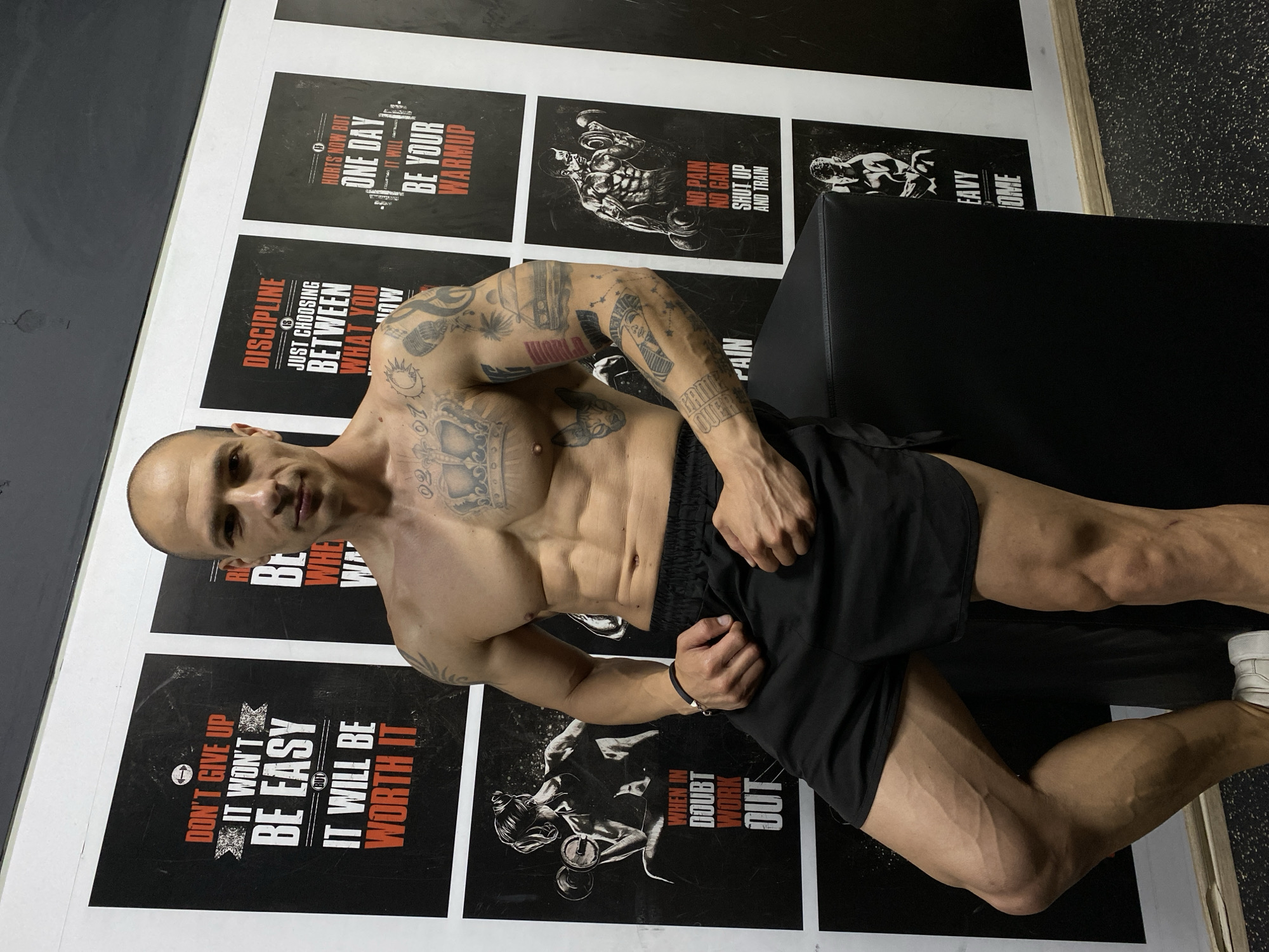 The image shows a muscular, bald man with tattoos posing in front of motivational posters in what appears to be a gym. He is wearing black shorts and white sneakers. The posters feature phrases like "Don't give up, it won't be easy, it will be worth it," "Discipline is just choosing between what you want now and what you want most," "Hurts now but one day it will be your warmup," "No pain no gain, shut up and train," and "When in doubt, work out." The background is a combination of black and white walls, and part of a black bench is visible. The location of Pleven, Bulgaria is not visually determinable from the image.