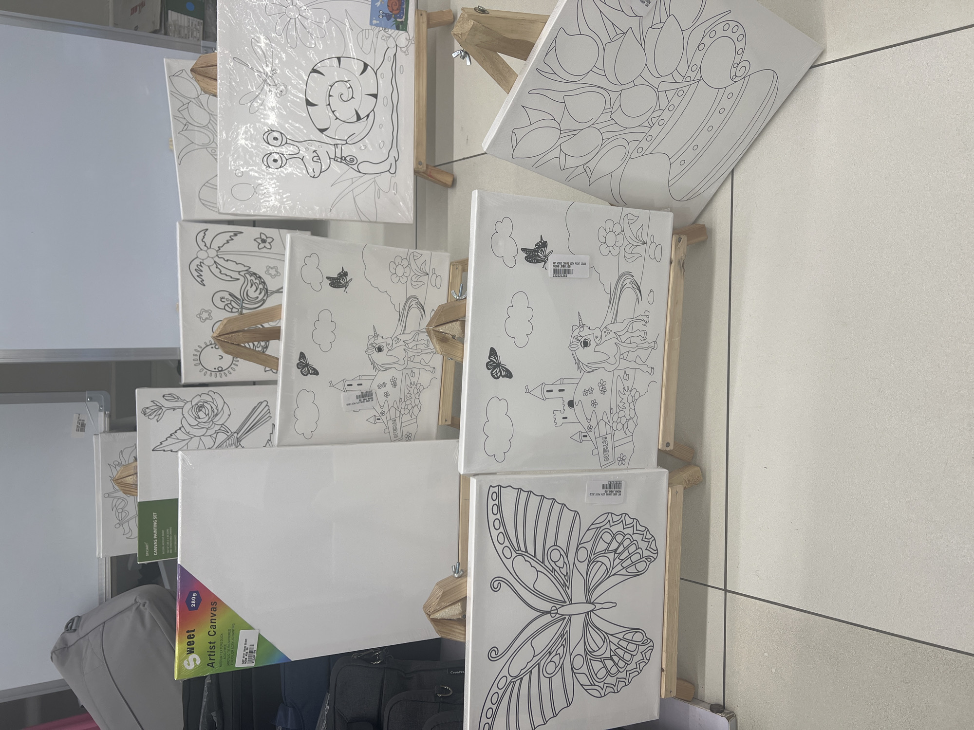 The image shows a display of various "Sweet Artist Canvas" and "Singart Canvas Painting Set" products, likely within a retail setting. The canvases are mounted on small wooden easels. The canvas designs include a snail, flowers, a beach scene with palm trees, unicorns, castles, and a butterfly. A plain white canvas is also visible. The canvases are arranged in a stacked manner, partially obscuring each other. There is luggage in the bottom left and bottom center of the image. The setting appears to be a store or market, possibly in Abuja, Nigeria.