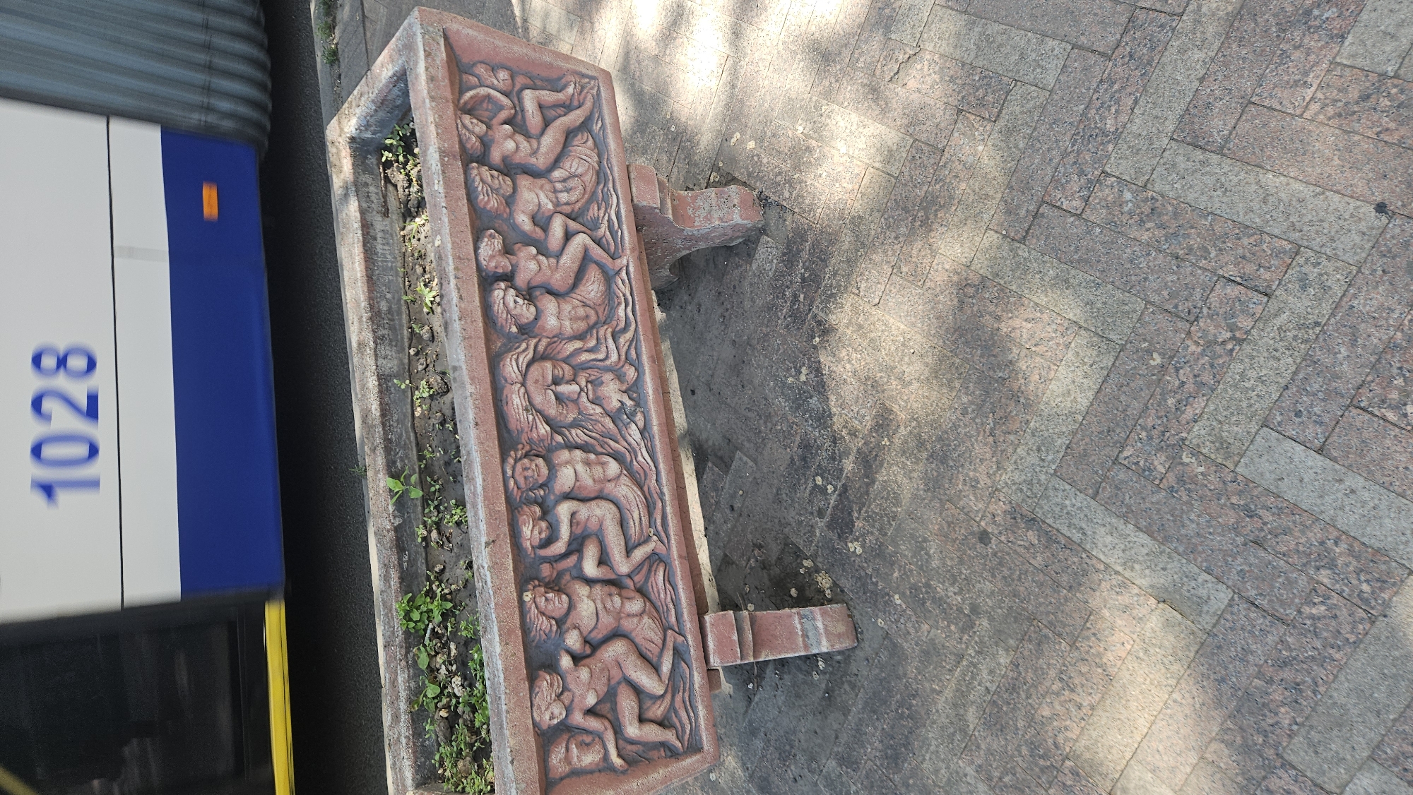 The image shows a long, rectangular concrete planter with classical-style bas-relief carvings of reclining human figures along its side, standing on a paved surface consisting of herringbone-patterned grey and pink granite tiles. Small green plants are visible inside the planter. To the left of the planter is the side of a bus or trolleybus with the number "1028" prominently displayed. The bus is predominantly white and blue, with a yellow stripe. The scene appears to be outdoors on a sunny day, with shadows cast on the paving. Based on the architectural style of the planter and available visual cues, the location is likely in a public space in Chișinău, Moldova.