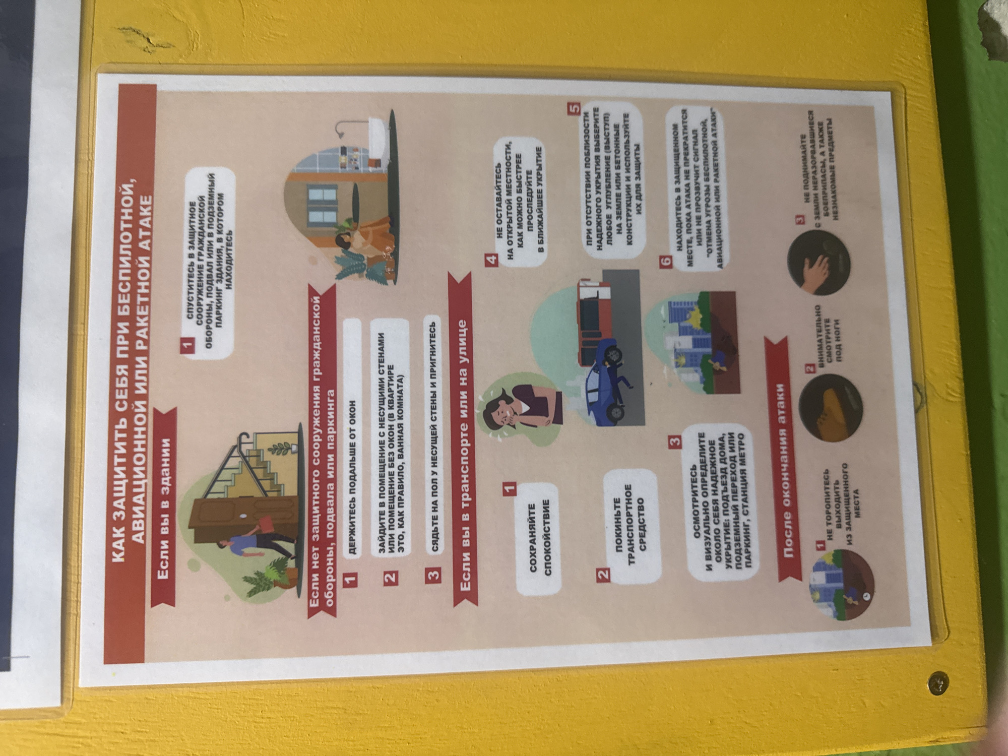 The image is a poster providing guidance on how to protect oneself during drone and rocket attacks. The poster has a yellow background and is mounted outdoors, likely on a building or a kiosk. It is written in Russian and divided into sections detailing actions to take if you are indoors, in a vehicle, or on the street, as well as post-attack procedures.
The "If you are indoors" section advises seeking shelter in a civil defense facility, basement, or underground parking, staying away from windows, entering a room with load-bearing walls or no windows (like a bathroom), and sitting on the floor near a load-bearing wall.
The "If you are in a vehicle or on the street" section suggests staying calm, leaving your vehicle, looking around for a safe shelter like a building entrance, underground passage, or subway station. It also advises against staying in open spaces and to choose a reliable underground shelter or concrete structures for protection. If in a protected area, it recommends staying there until the threat subsides or an all-clear signal is given.
The "After the attack" section instructs not to rush to leave the shelter, to carefully check the ground for unexploded ordnance, and not to touch unfamiliar objects.
The poster features illustrative drawings depicting the advice given in each section, showing people taking shelter, vehicles, and buildings. The overall color scheme is dominated by red and beige for the text and graphics, against a plain white background within the poster frame. The yellow exterior surface suggests it's an outdoor information board.
It is impossible to determine the specific location in the city of Saratov, Russia, from the image alone.